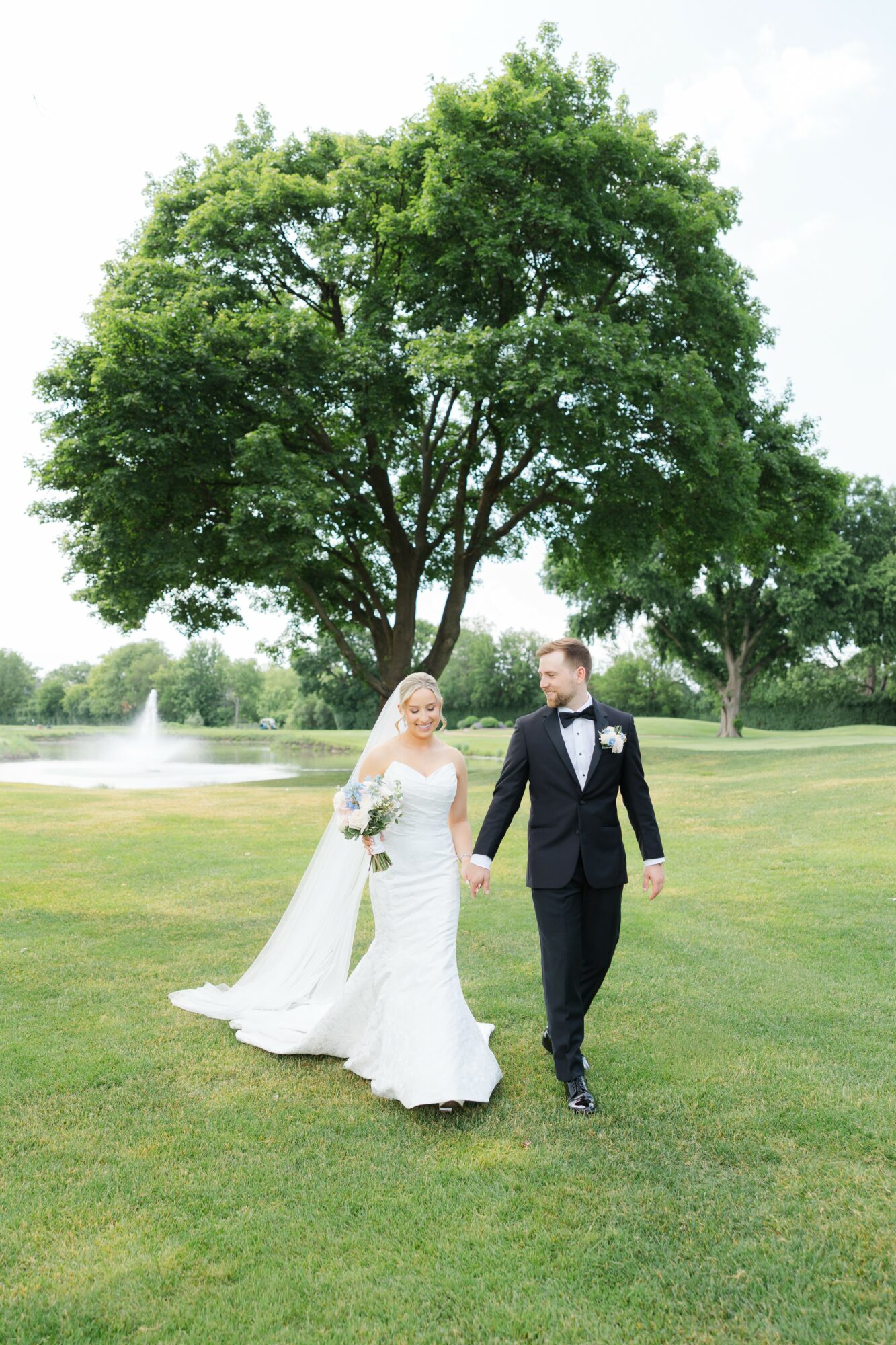 Bride and groom walking on grass near a large tree, with a fountain in the background, outdoors on a sunny day.