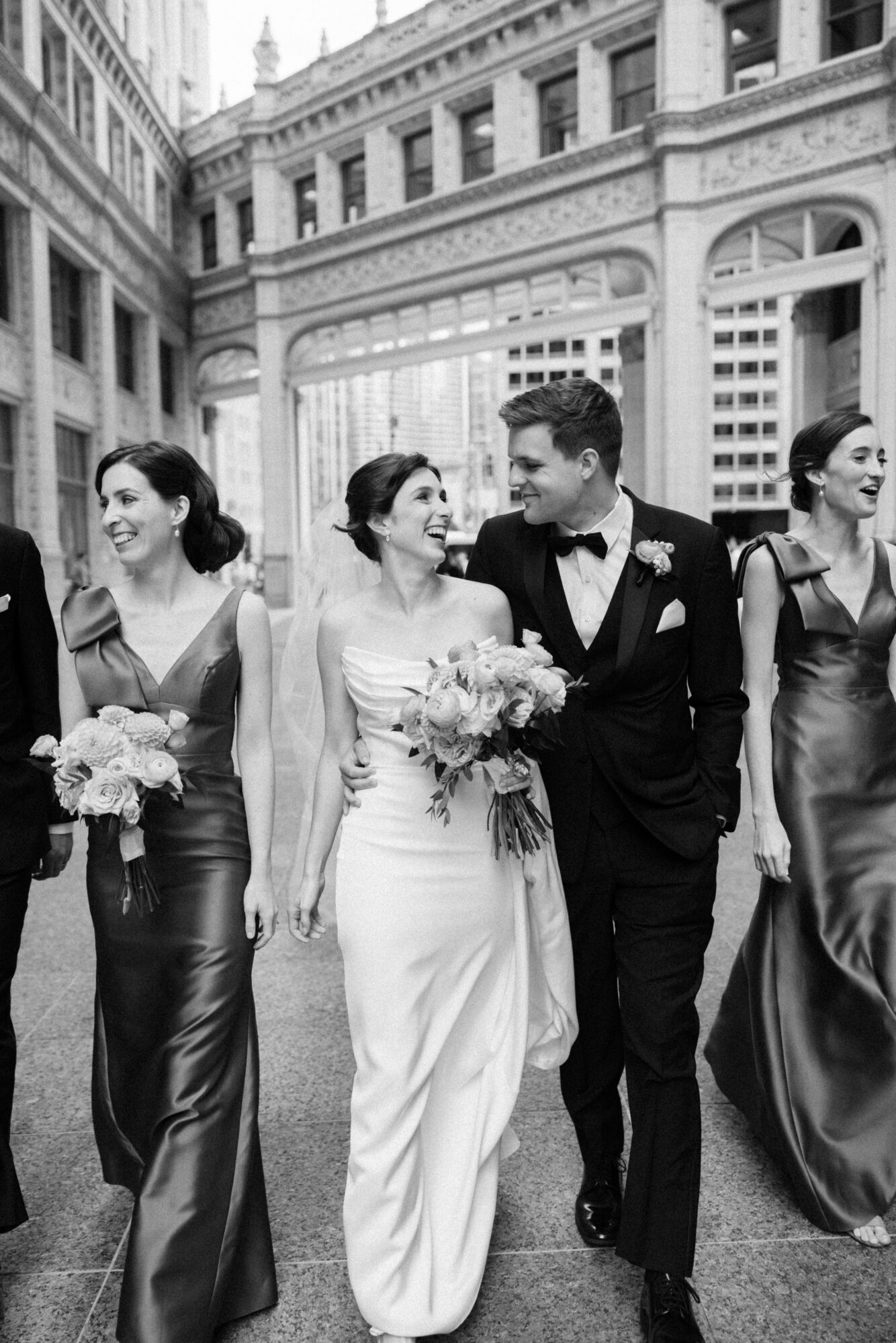 Group of people in formal attire walking outdoors, smiling, with a historic building in the background.
