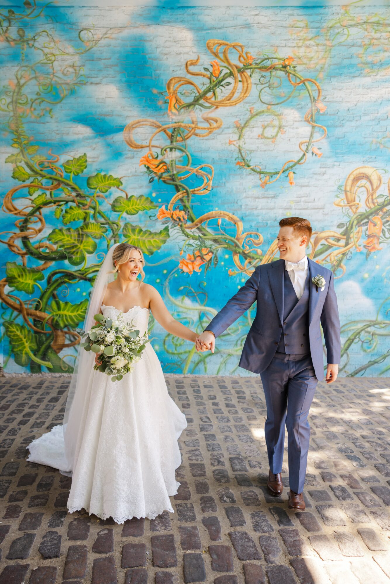 Bride and groom holding hands, walking on cobblestone, with a colorful mural of trees and swirling patterns in the background.