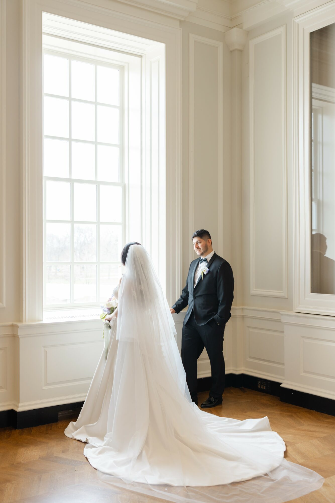 Bride and groom holding hands near large window in elegant room, bride in white gown with train, groom in dark suit.