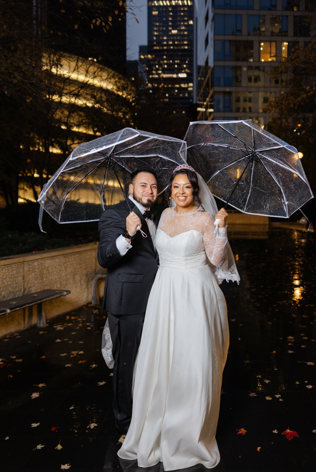 Bride and groom holding transparent umbrellas outdoors at night, city lights in background, bride in white gown, groom in dark suit.