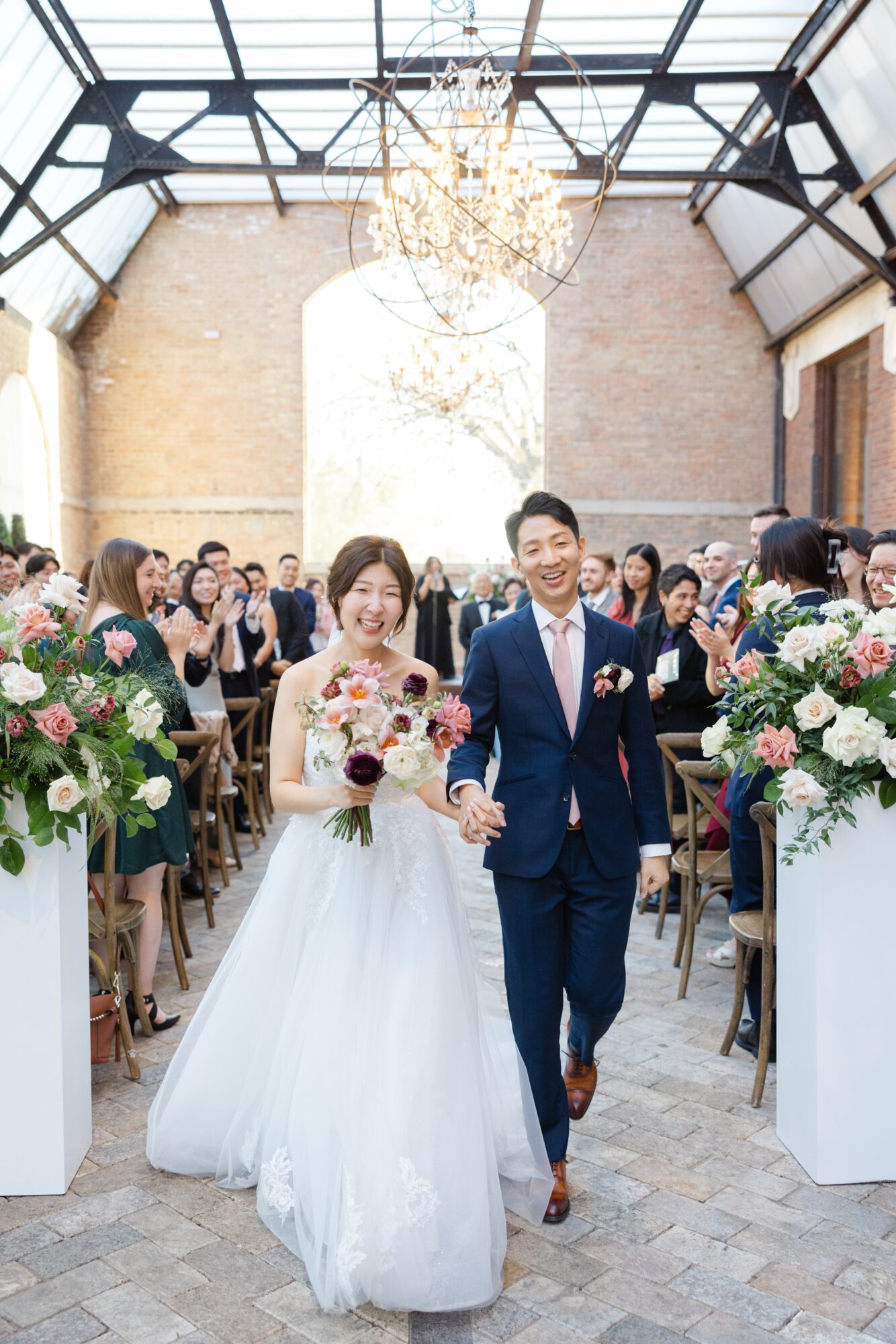 Bride and groom walking down aisle holding hands, guests seated on sides, floral arrangements, chandelier overhead, bright natural light.