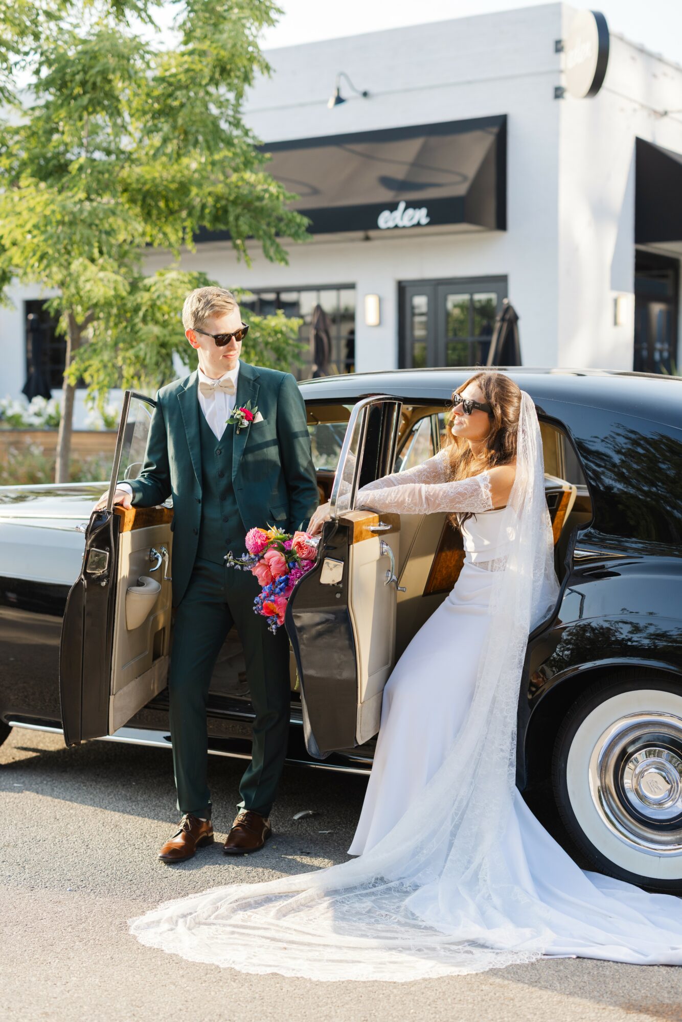 Bride in white dress with train and sunglasses, holding a bouquet, exits a black limousine, groom in suit stands nearby outdoors.