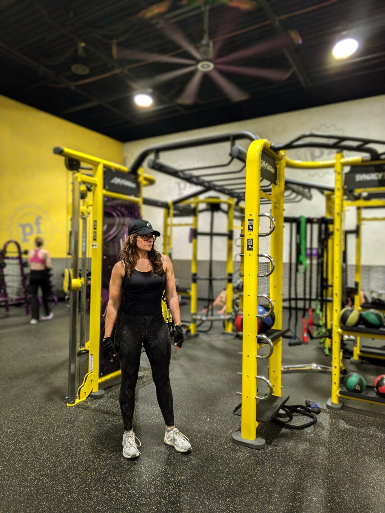 Woman in black workout clothes and cap standing in gym with yellow and black exercise equipment, black ceiling, and other people in background.