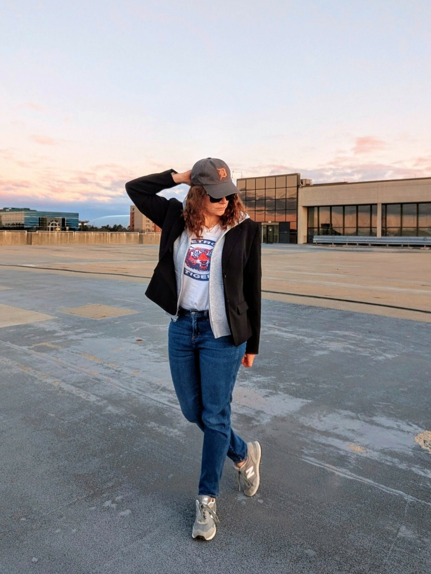 Person walking outdoors on a paved surface, wearing a cap, jacket, jeans, and sneakers, with modern buildings in the background.
