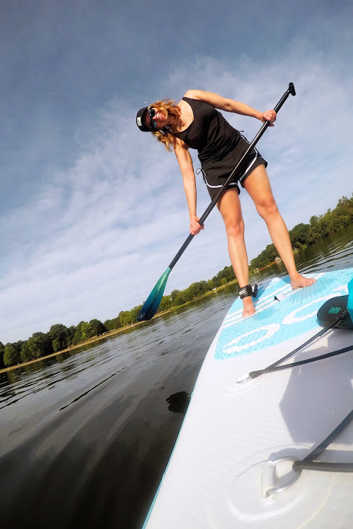 Person standing on paddleboard, holding paddle, leaning forward, water and sky in background.