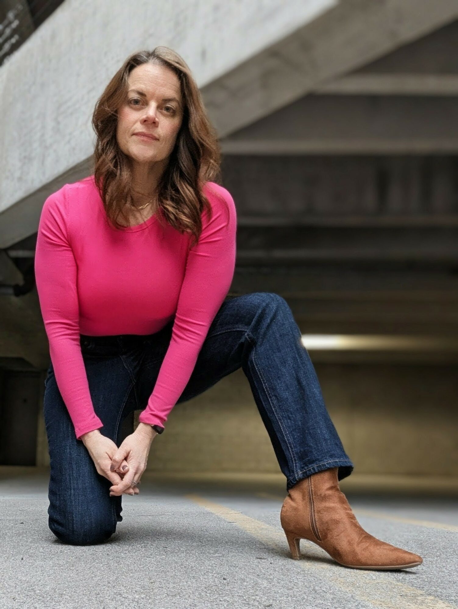 Woman crouching outdoors, wearing a pink top, jeans, and brown ankle boots, under a concrete structure.