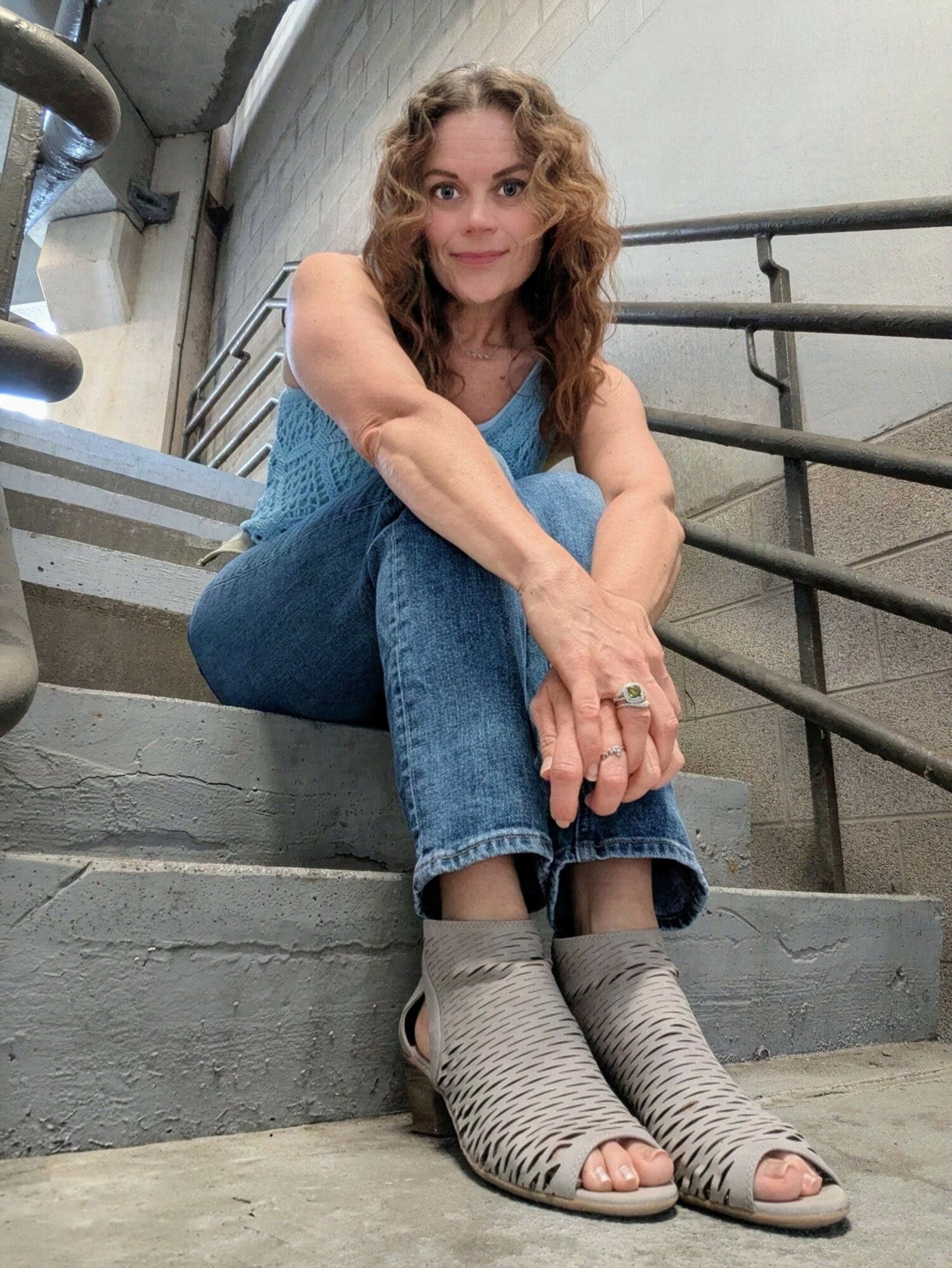Woman with curly hair sitting on stairs, wearing jeans and open-toe shoes, smiling at camera.