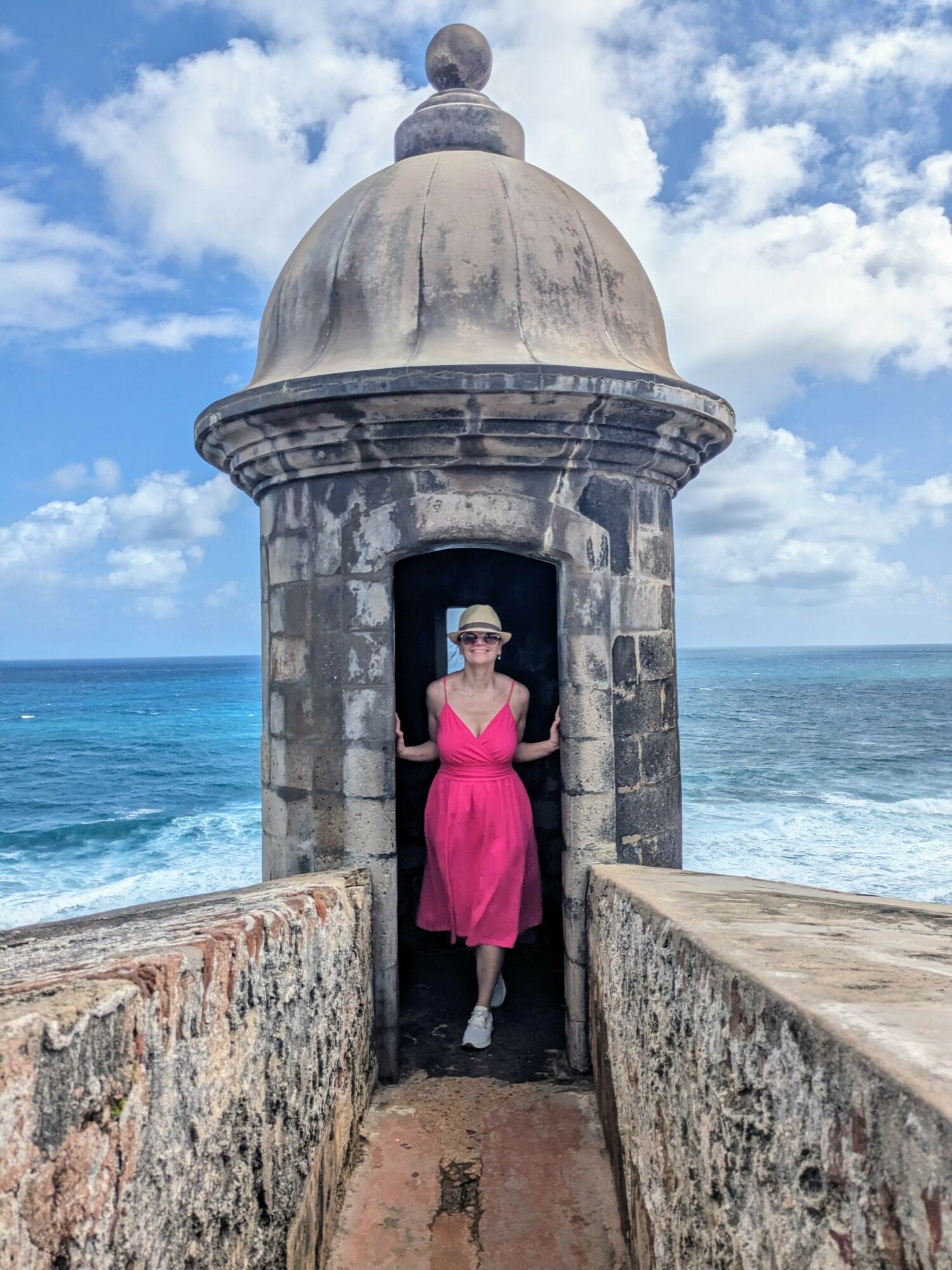 Woman in pink dress and hat standing inside a small stone structure by the sea, with a domed roof and blue sky.