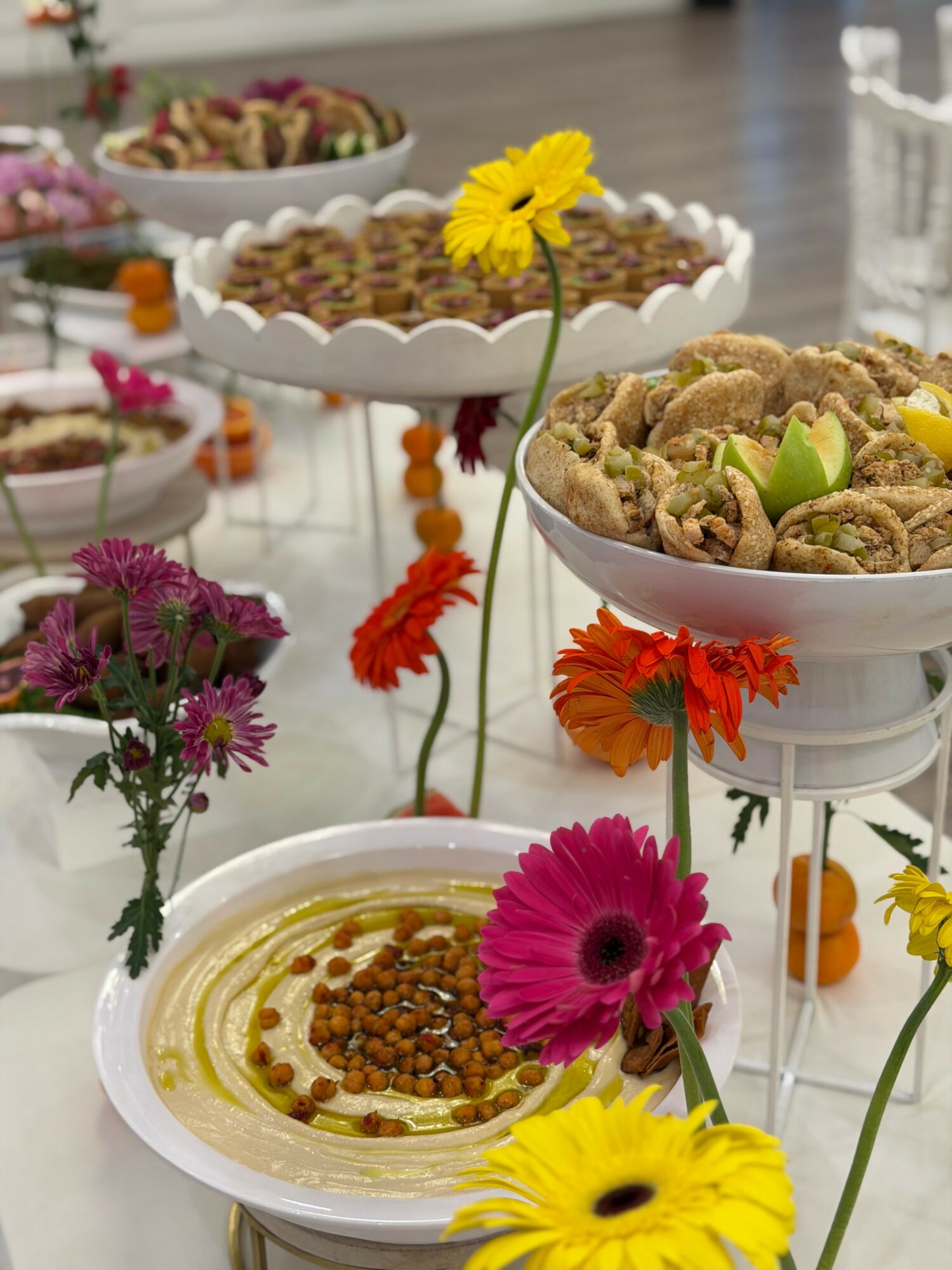 Table with bowls of food and colorful flowers, including a yellow flower in the center, in a bright setting.