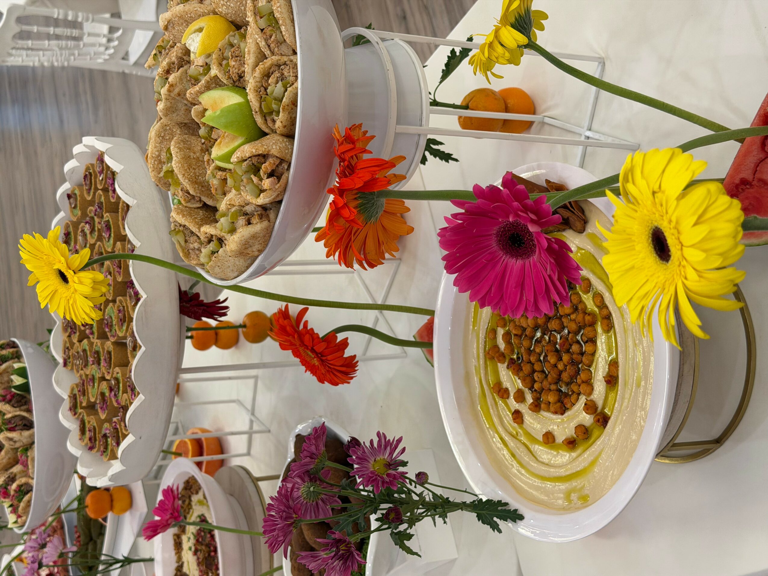 Table with plates of food and colorful flowers, including yellow, pink, and orange blossoms, on a white surface.