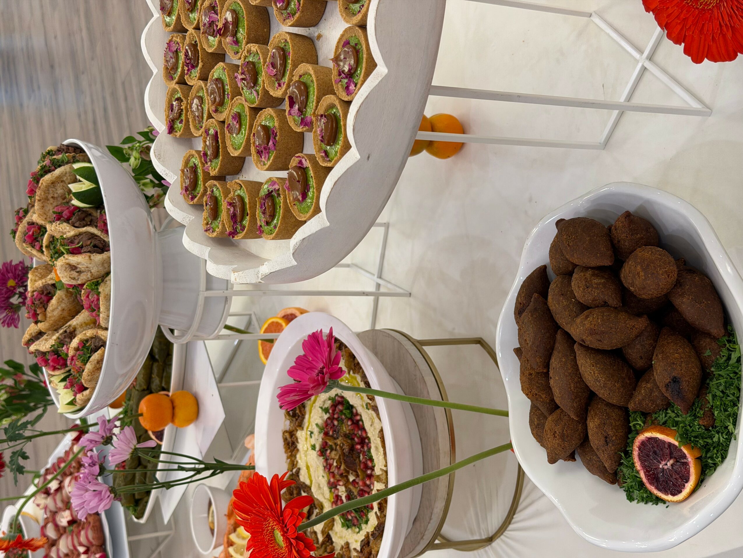 Assorted desserts and flowers on white tables, including chocolate truffles, rolled pastries, and a cake with pink flowers.
