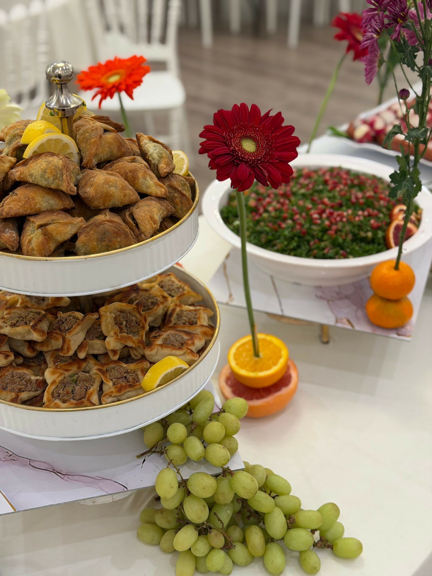 Plate of fried snacks, grapes, and decorative flowers on a table, with blurred chairs in background.