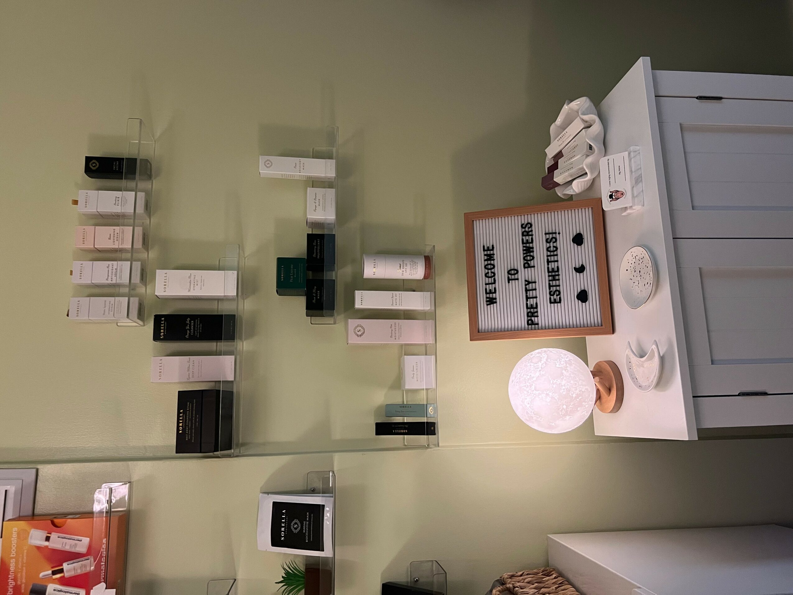 Wall with organized nail polish bottles, a sign, and a lamp on a white table, with a laundry machine nearby.