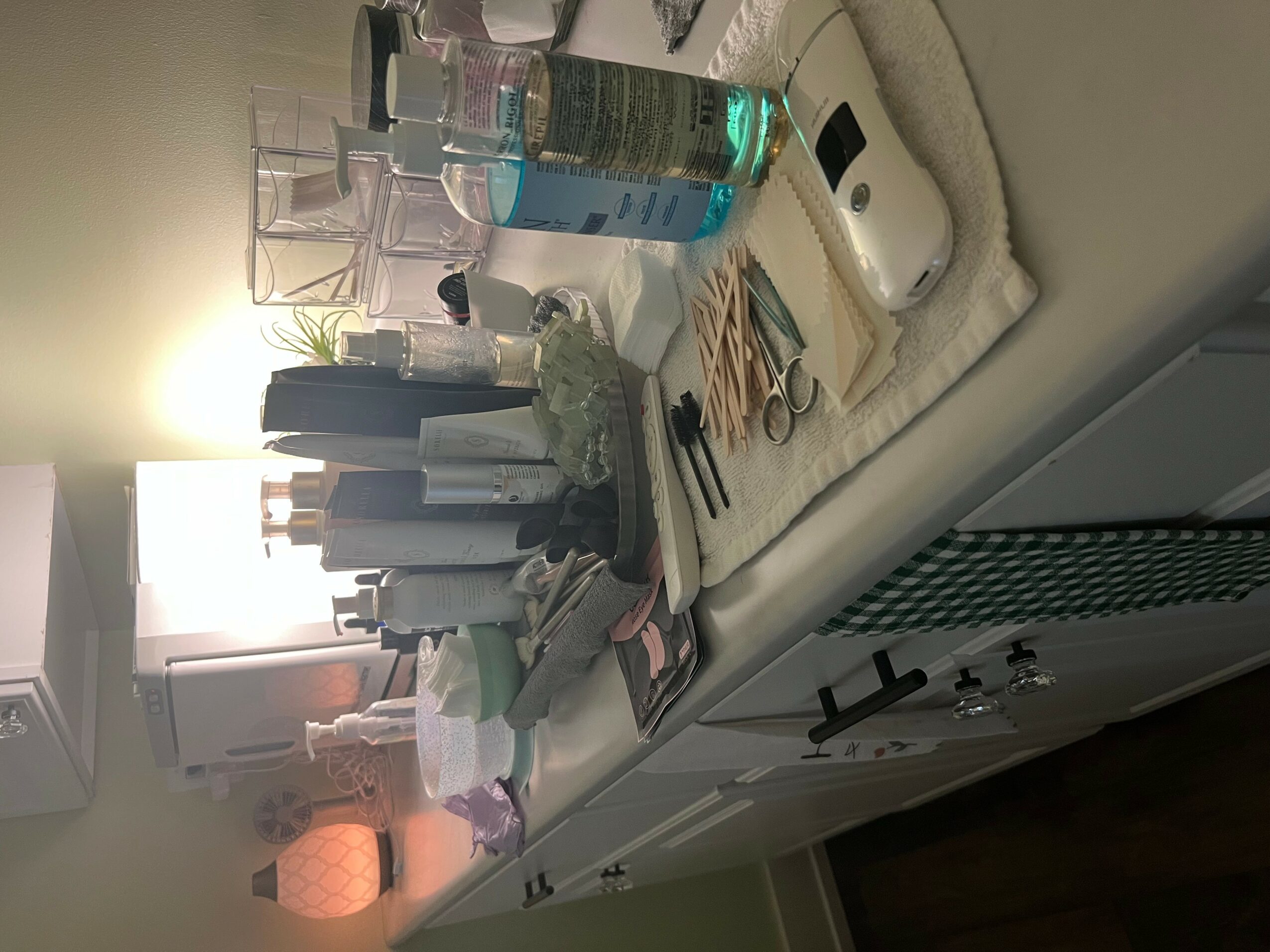Cluttered countertop with bottles, papers, and scissors, next to a white device and a beige cushion, with a bright background.