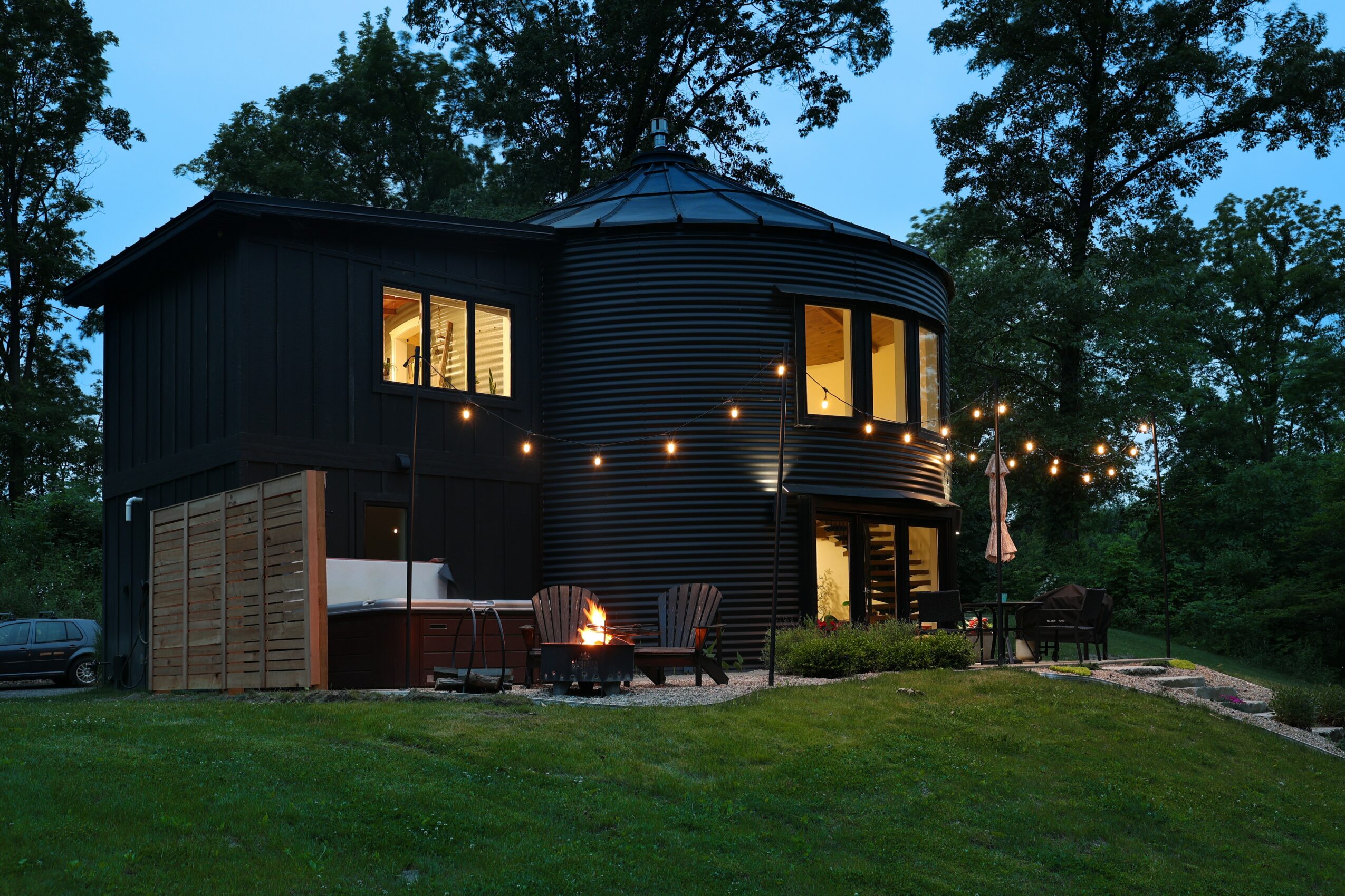 Black house with string lights, outdoor seating, and fire pit on a grassy yard surrounded by trees at dusk.