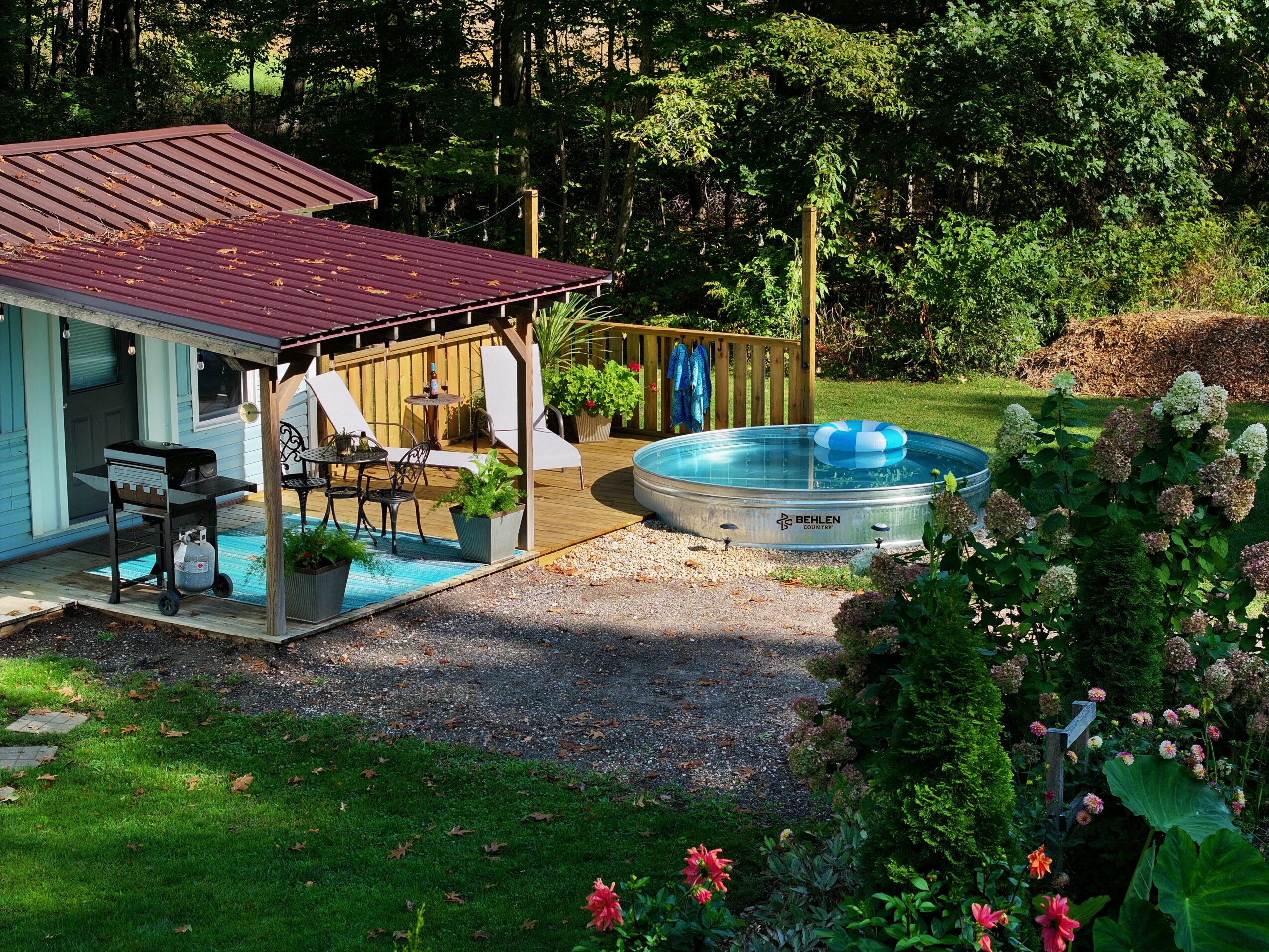 Backyard with a small pool, wooden deck, and garden, surrounded by trees and greenery.