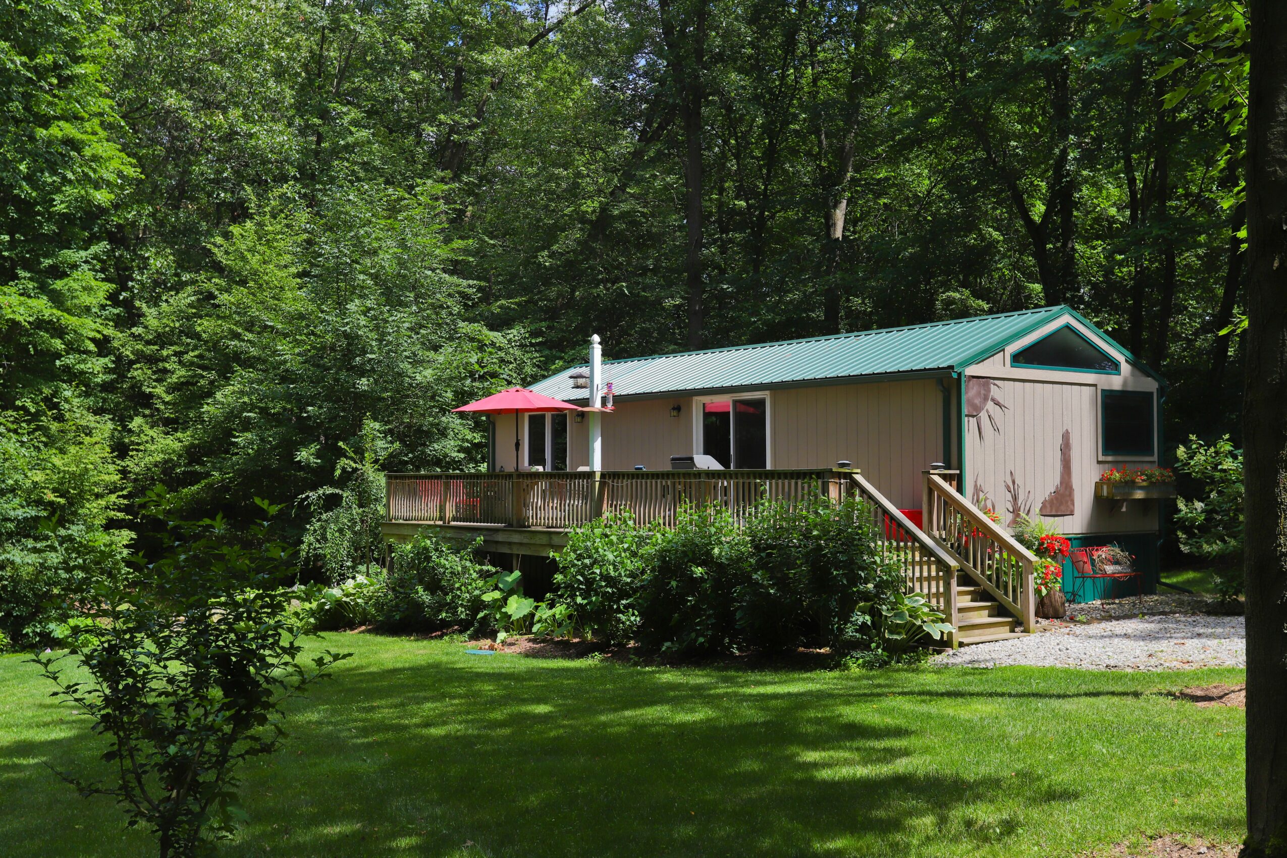 Single-story house with a deck, surrounded by trees and a grassy yard, under a clear sky.