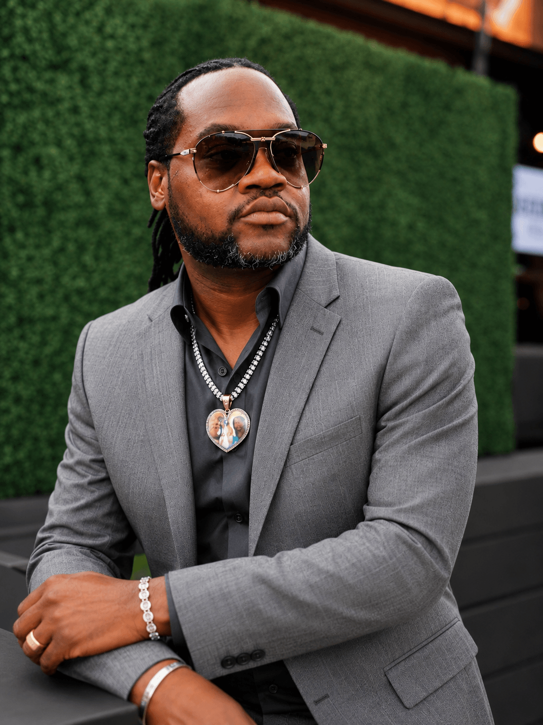 Man with sunglasses, beard, and long hair in a gray suit and black shirt, standing outdoors with green hedge background.