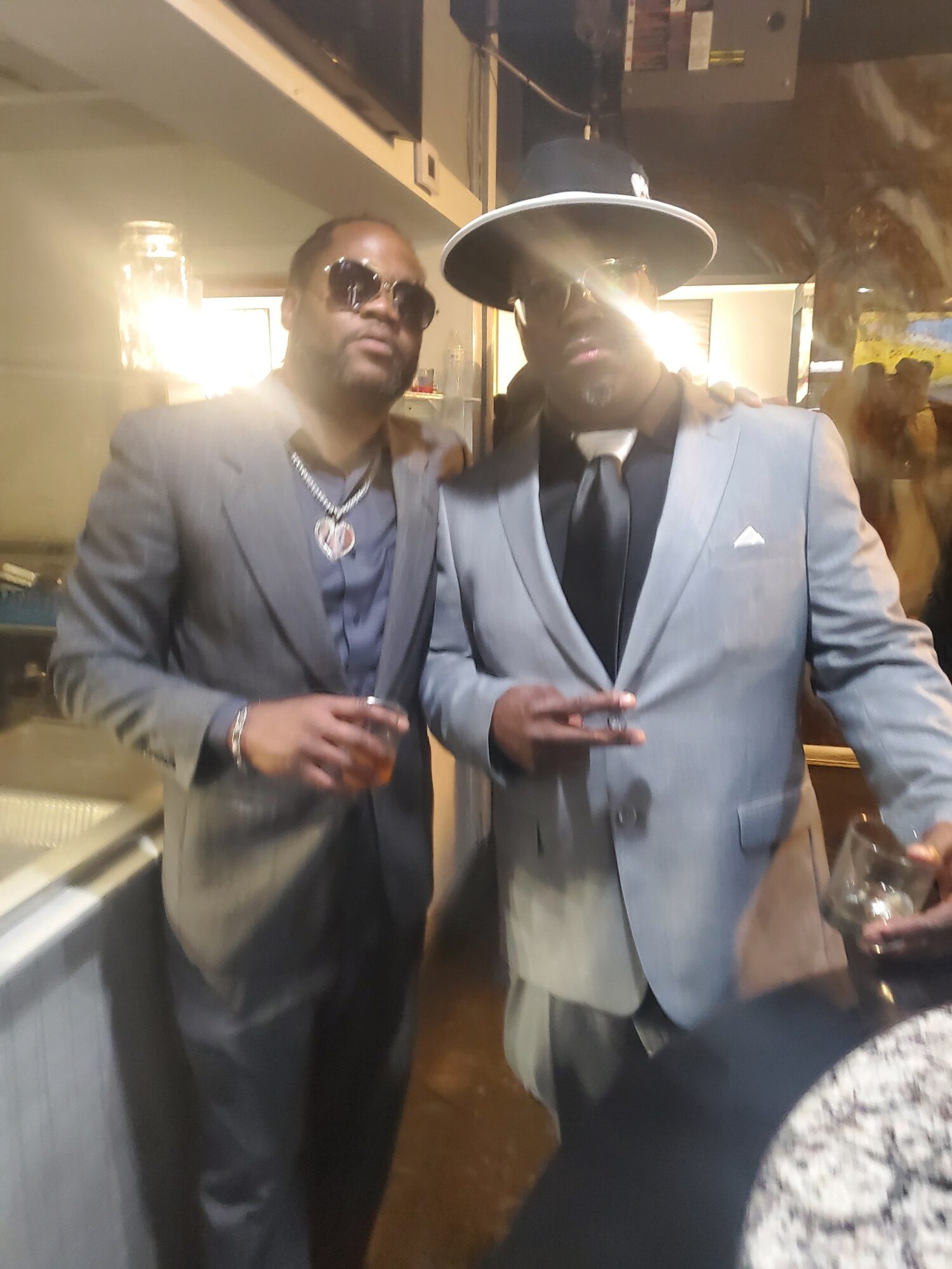 Two men in suits and sunglasses standing indoors, one holding a drink, with warm lighting and blurred background.