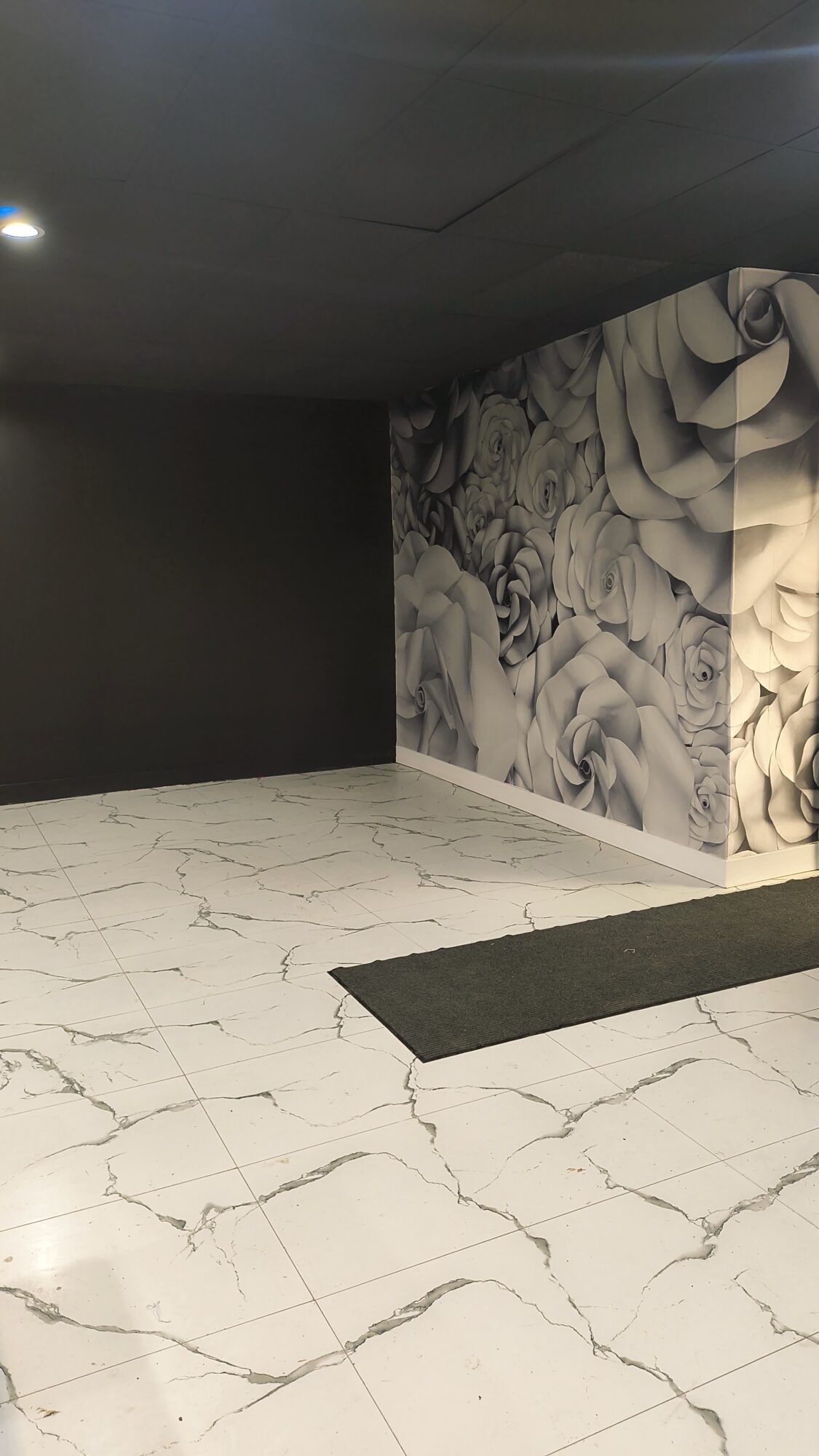 Corner of a room with black and patterned walls, marble floor, and a black mat, ceiling with a light fixture.