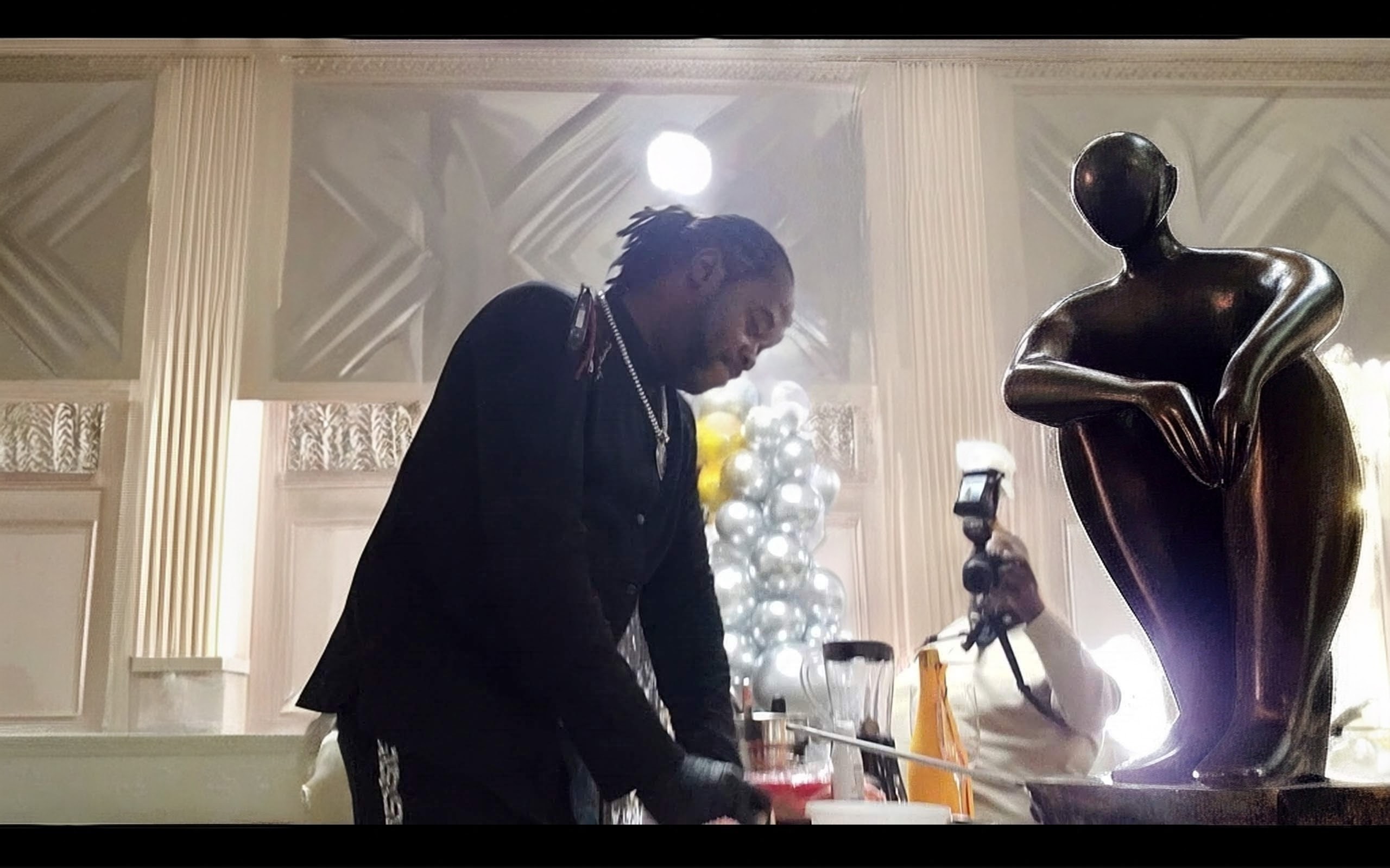 Person in dark clothing leaning over a table with a sculpture nearby, decorated room with columns and a Christmas tree in background.