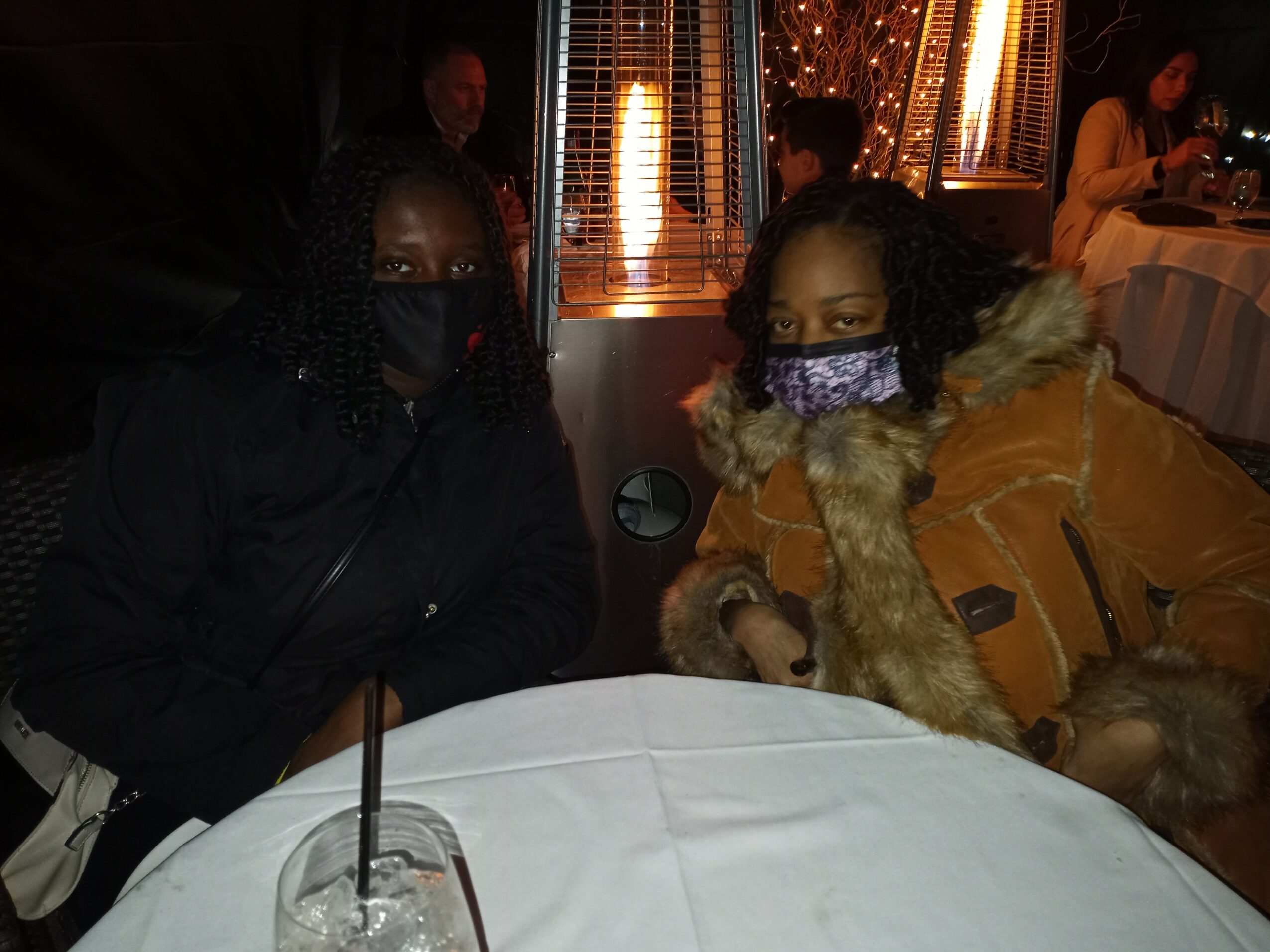 Two women wearing masks sit at a table in a dimly lit restaurant with warm lighting and other diners in the background.