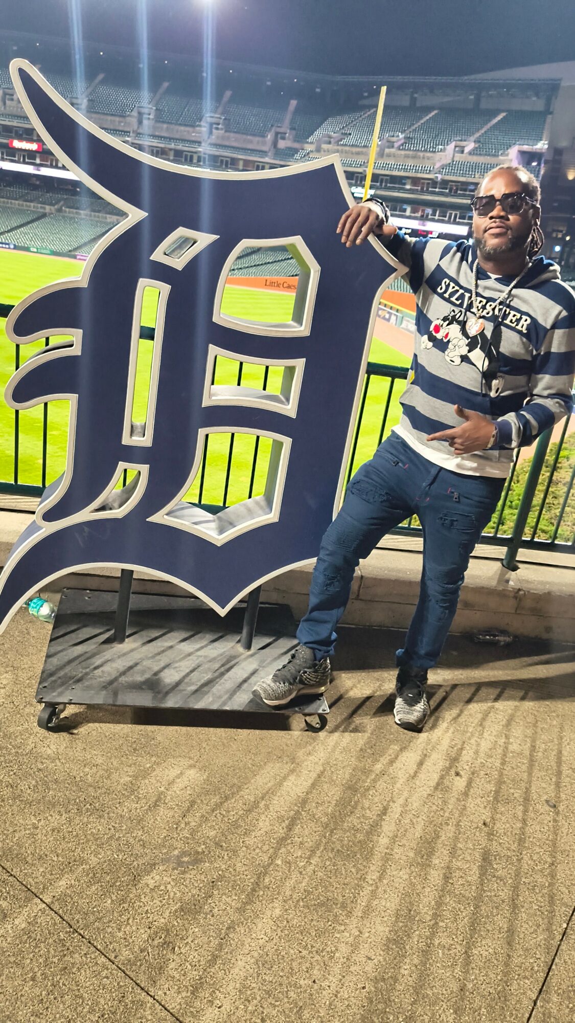 Person standing next to a large Detroit Tigers logo at a stadium, holding a yellow baseball bat, wearing sunglasses and a striped shirt.