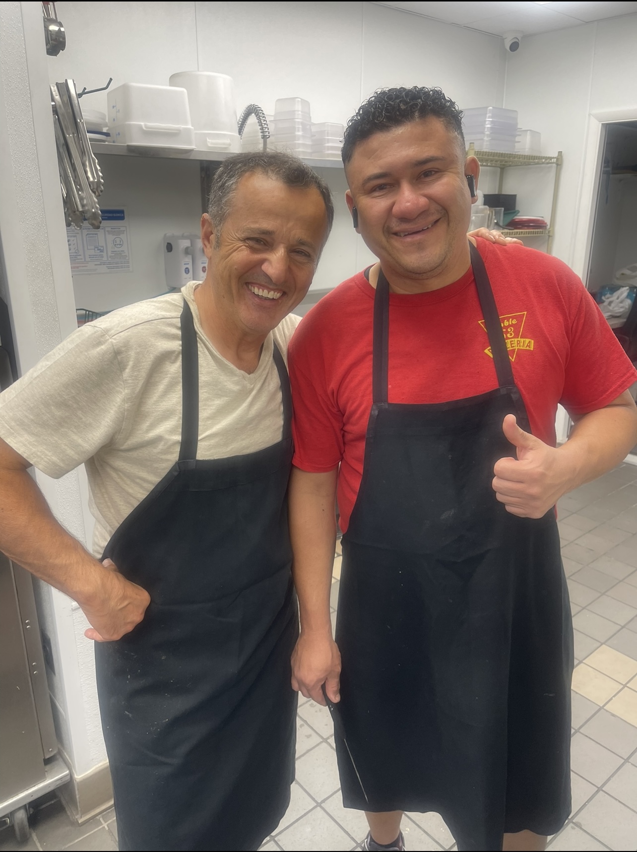 Two smiling men wearing aprons in a kitchen, one giving a thumbs-up, standing close together.