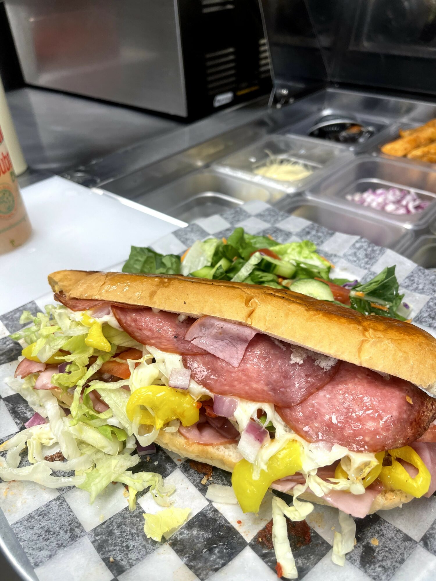 Sub sandwich with lettuce, ham, salami, and yellow peppers on a checkered paper, with salad and condiments in the background.