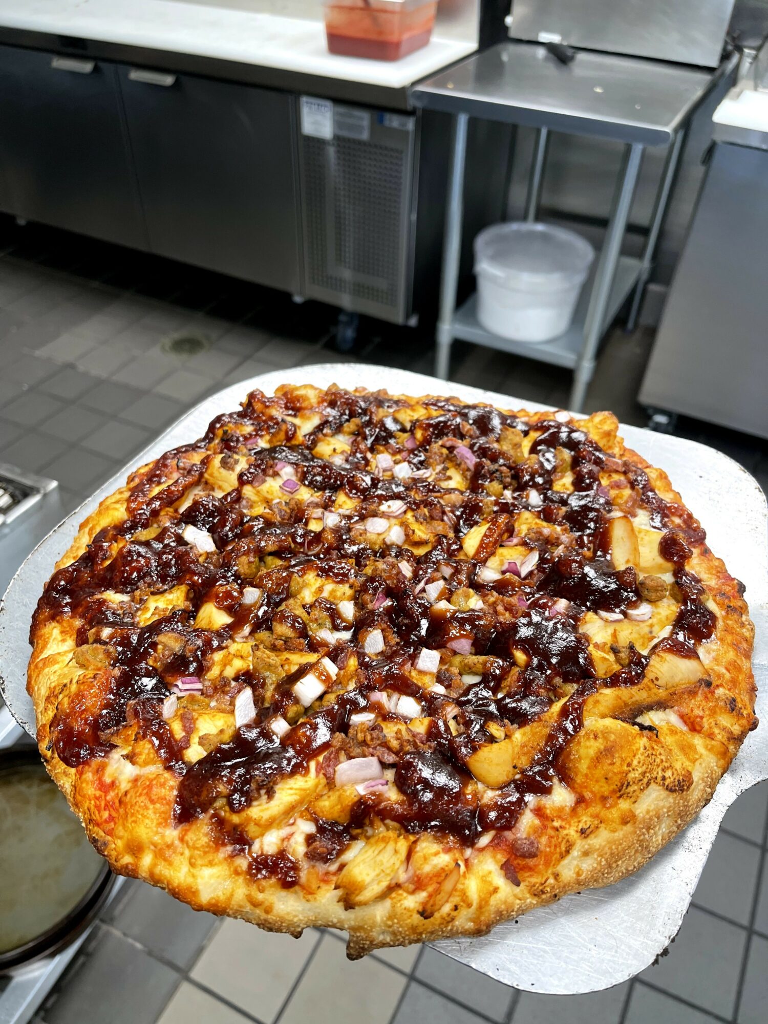 Large pizza with toppings, held over a kitchen counter, with a commercial kitchen background.