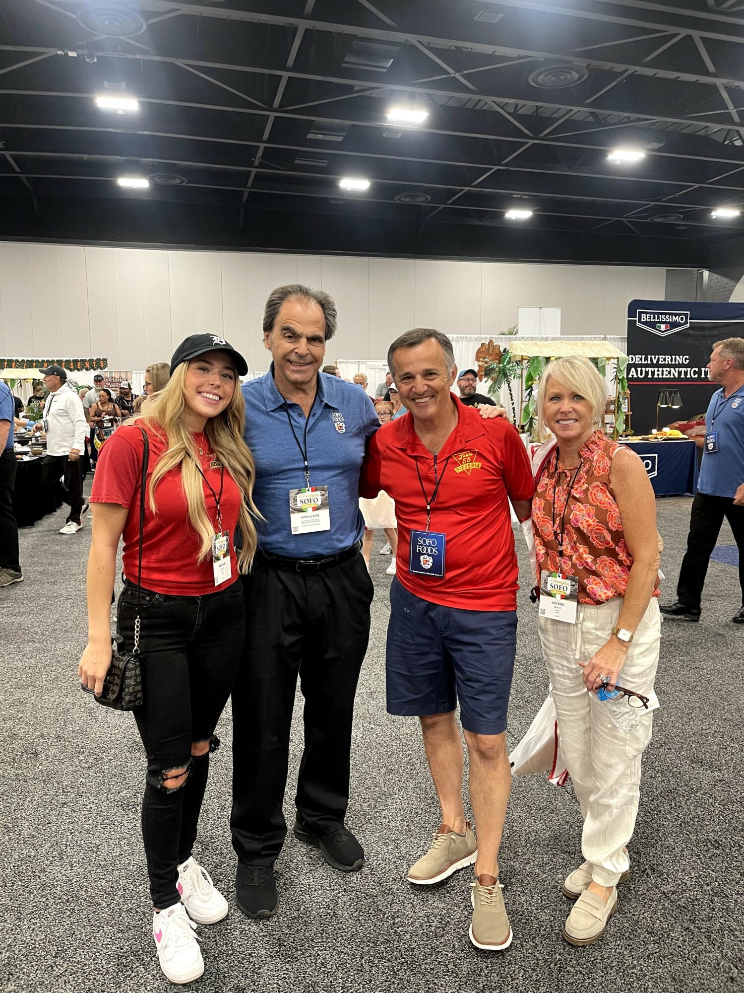 Four people standing together indoors at an event, smiling, with booths and displays in the background.