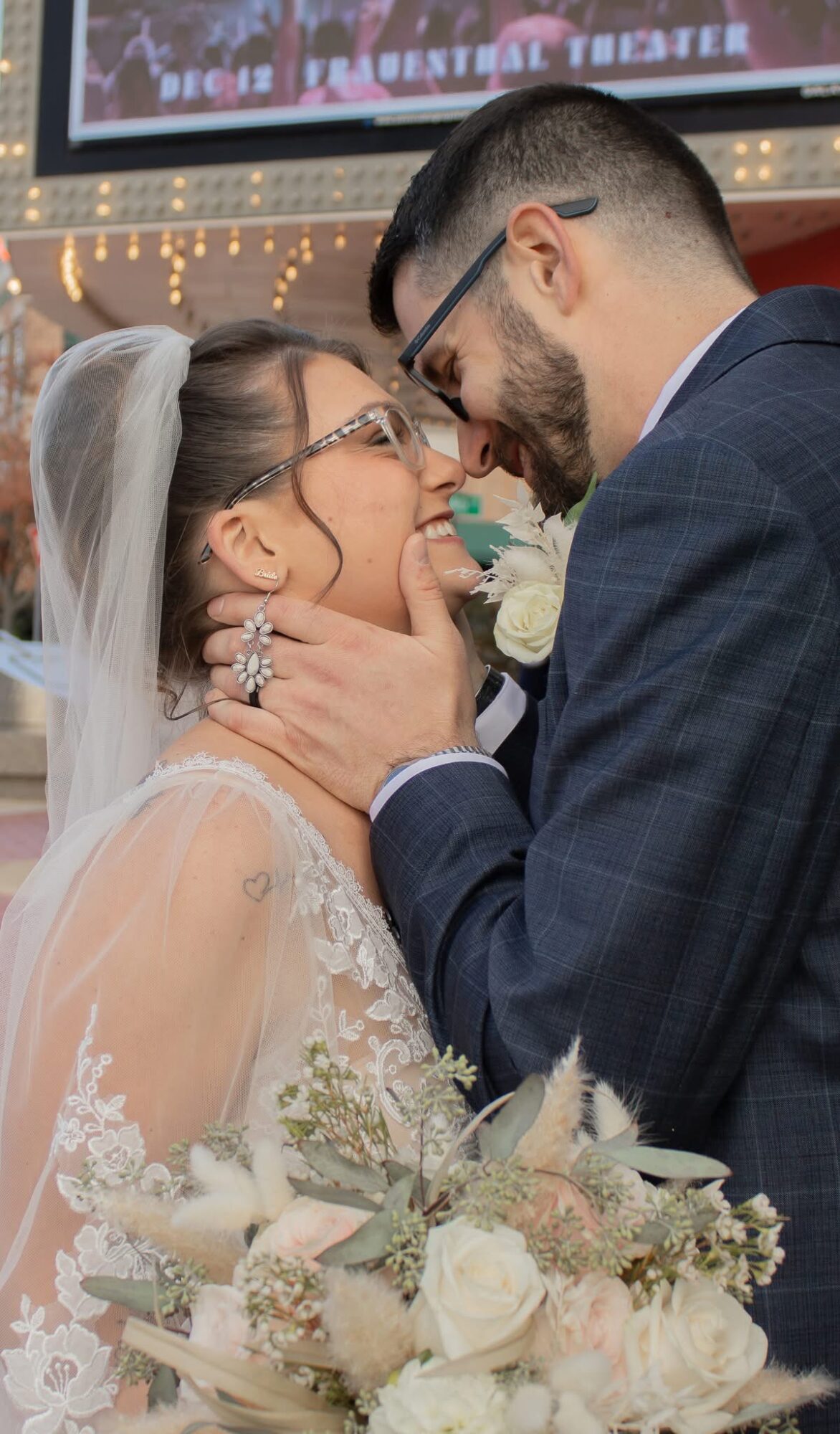 Bride and groom face each other, smiling, with the groom holding the bride's face gently, wedding flowers in foreground.