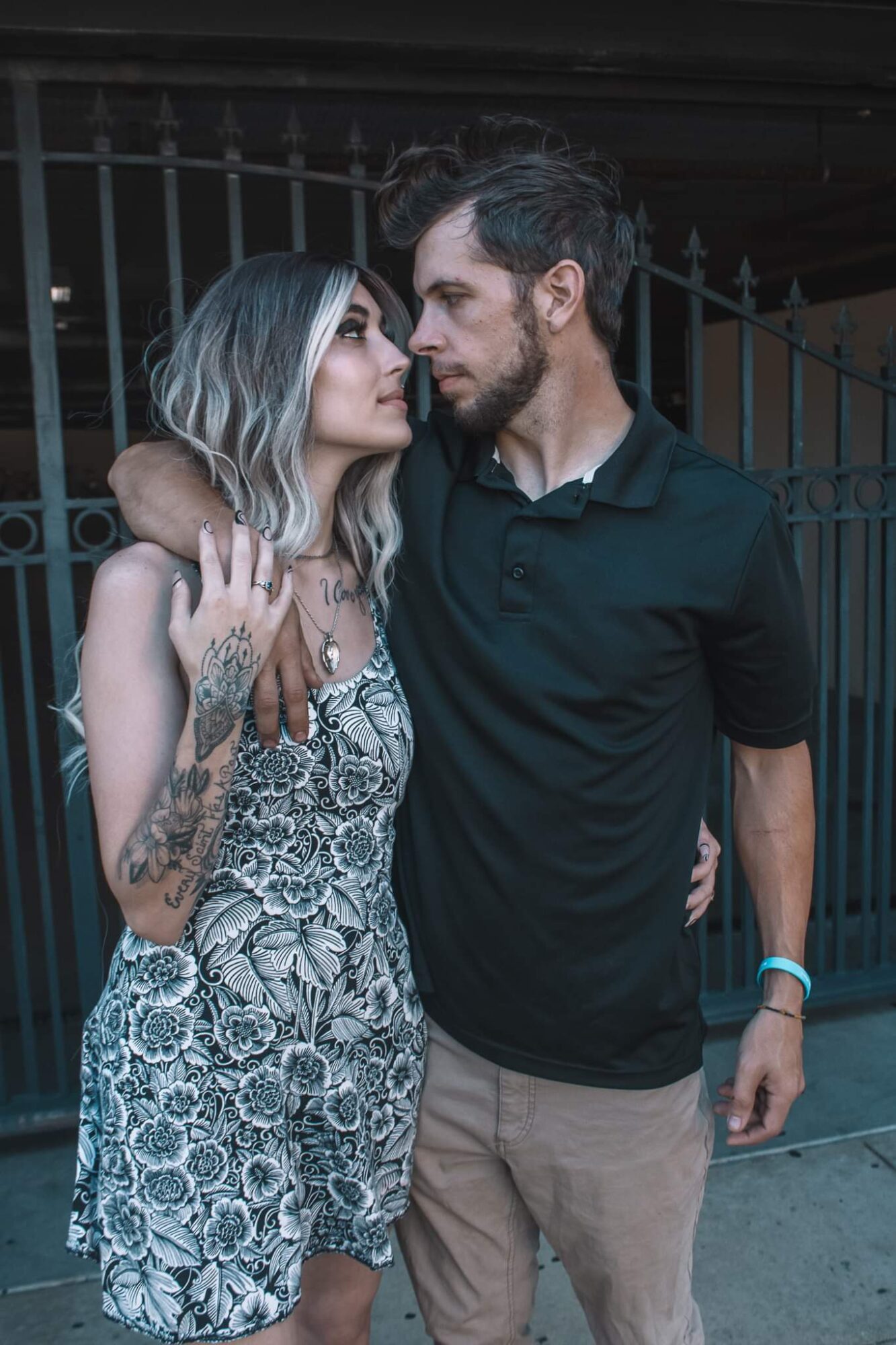 A man and woman stand close together, gazing into each other's eyes, with a fence in the background.