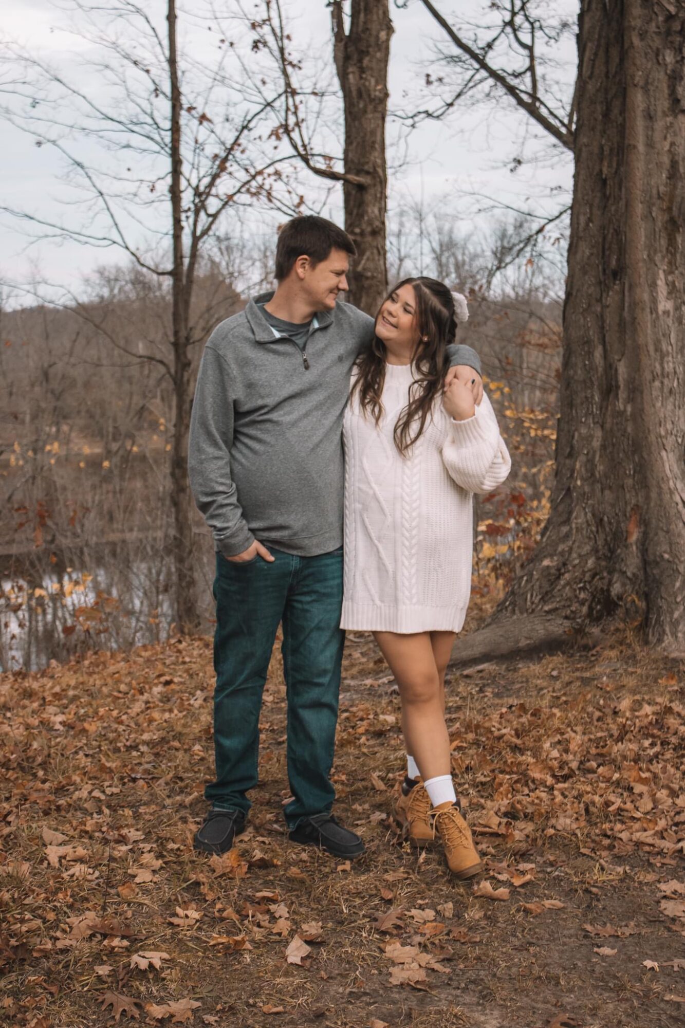 A young man and woman stand close together outdoors in a wooded area with fallen leaves, smiling at each other.