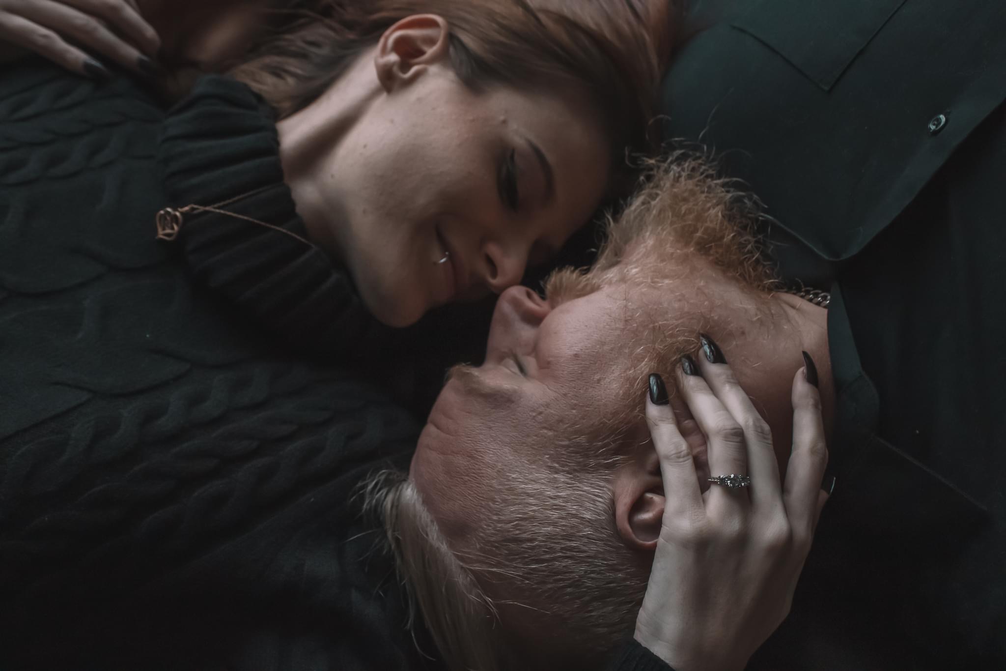 Two people lying close together, one with hand on the other's face, eyes closed, in an intimate moment.