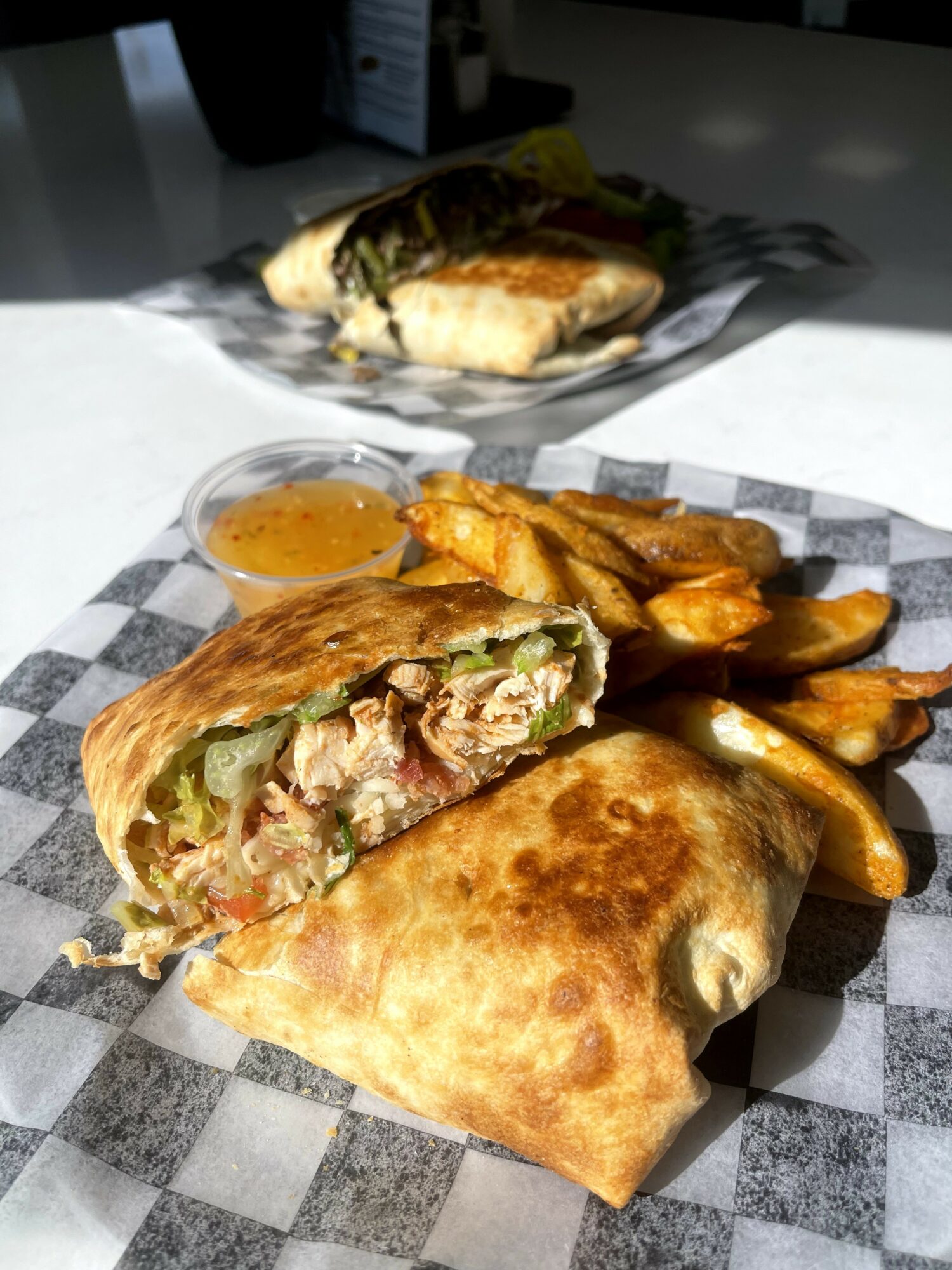 Fried spring roll cut in half with chicken, vegetables, and sauce, served with potato wedges on checkered paper.