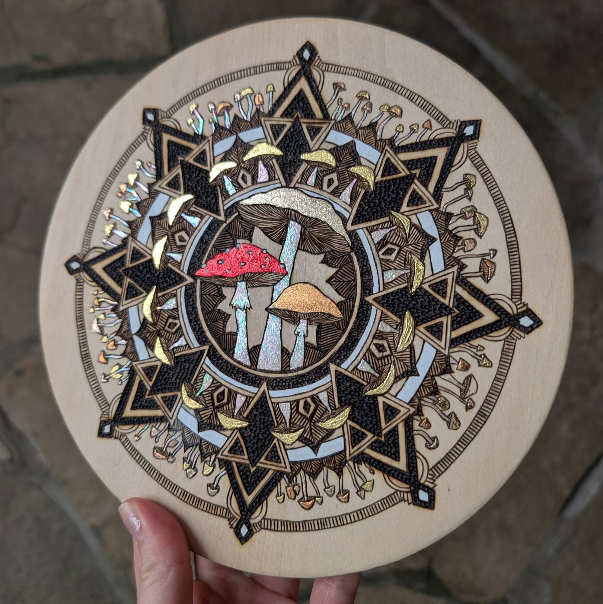 Decorative circular artwork with geometric patterns and three colorful mushrooms in the center, held by a person's hand.