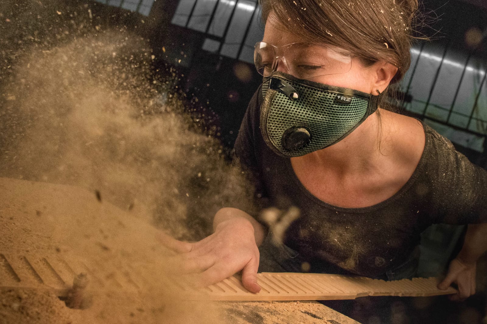 Person wearing a mask, leaning over a surface, with dust or particles flying around, indoors with a dark background.