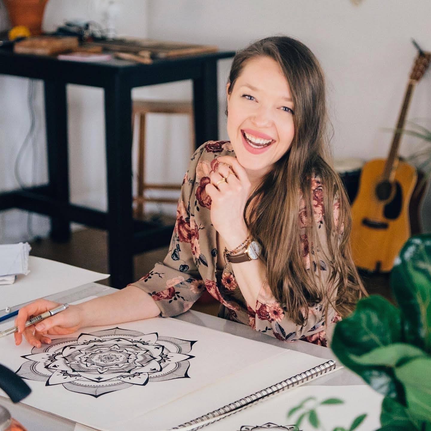 Woman with long hair smiling and holding a pen, sitting at a table with drawings, in a room with a guitar and furniture.