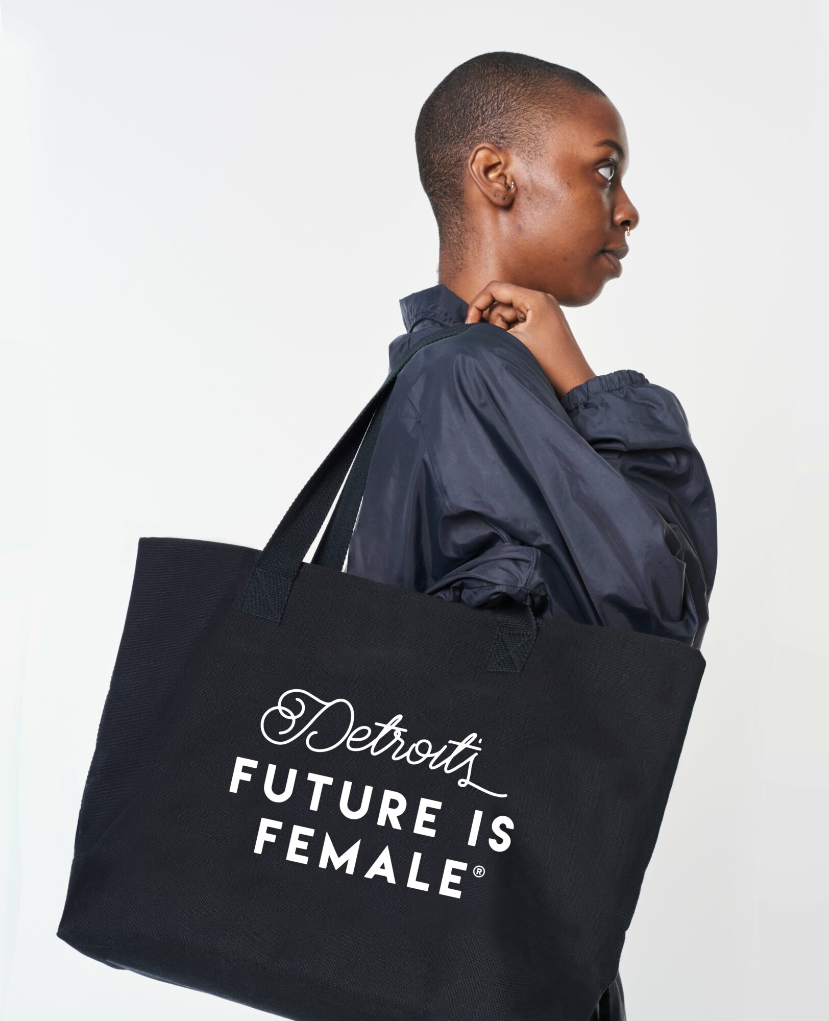 Side profile of a woman with short hair holding a black tote bag with white text, against a plain white background.