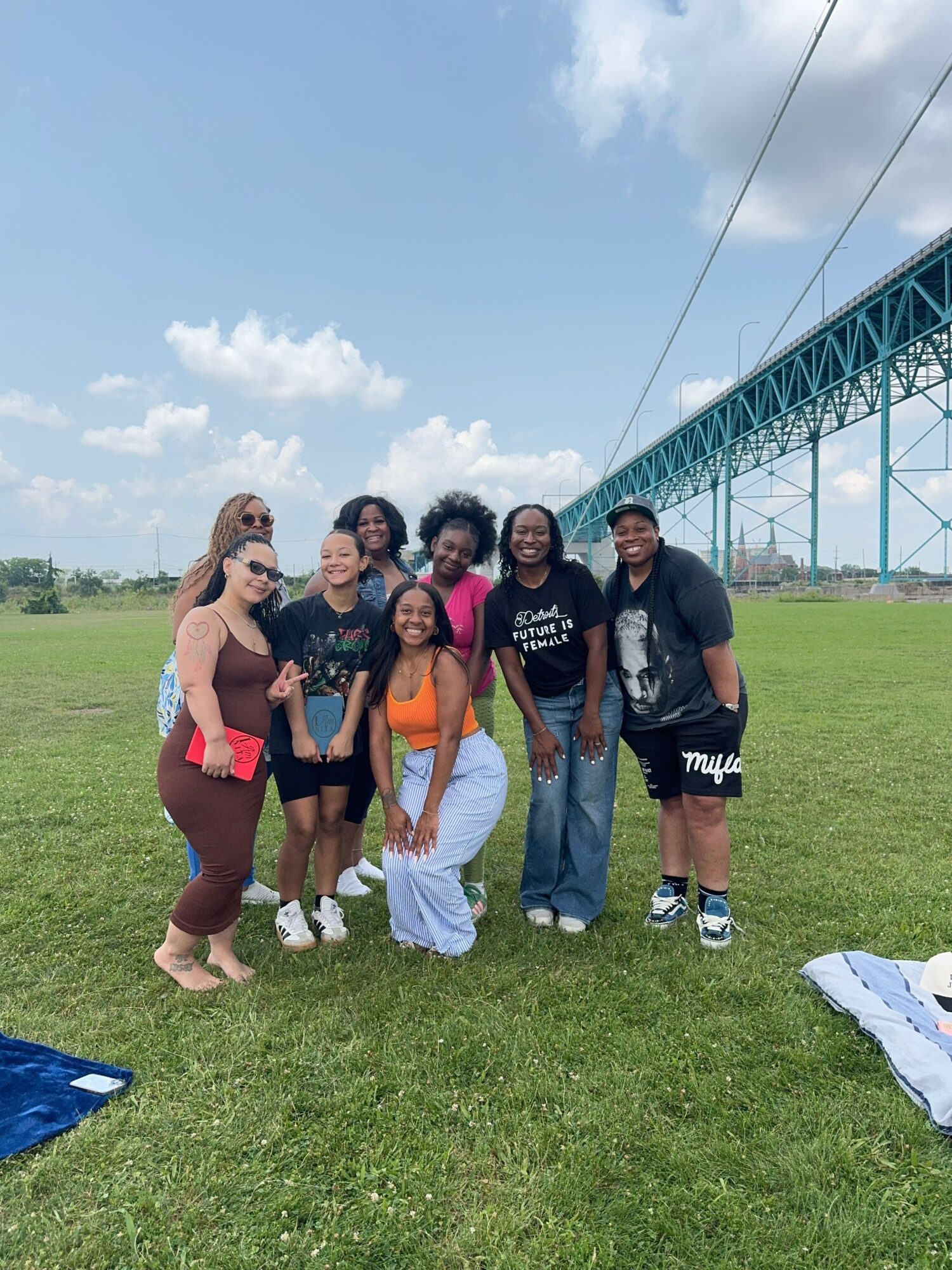 Group of six people standing on grass near a bridge, with a blue sky and clouds in the background.