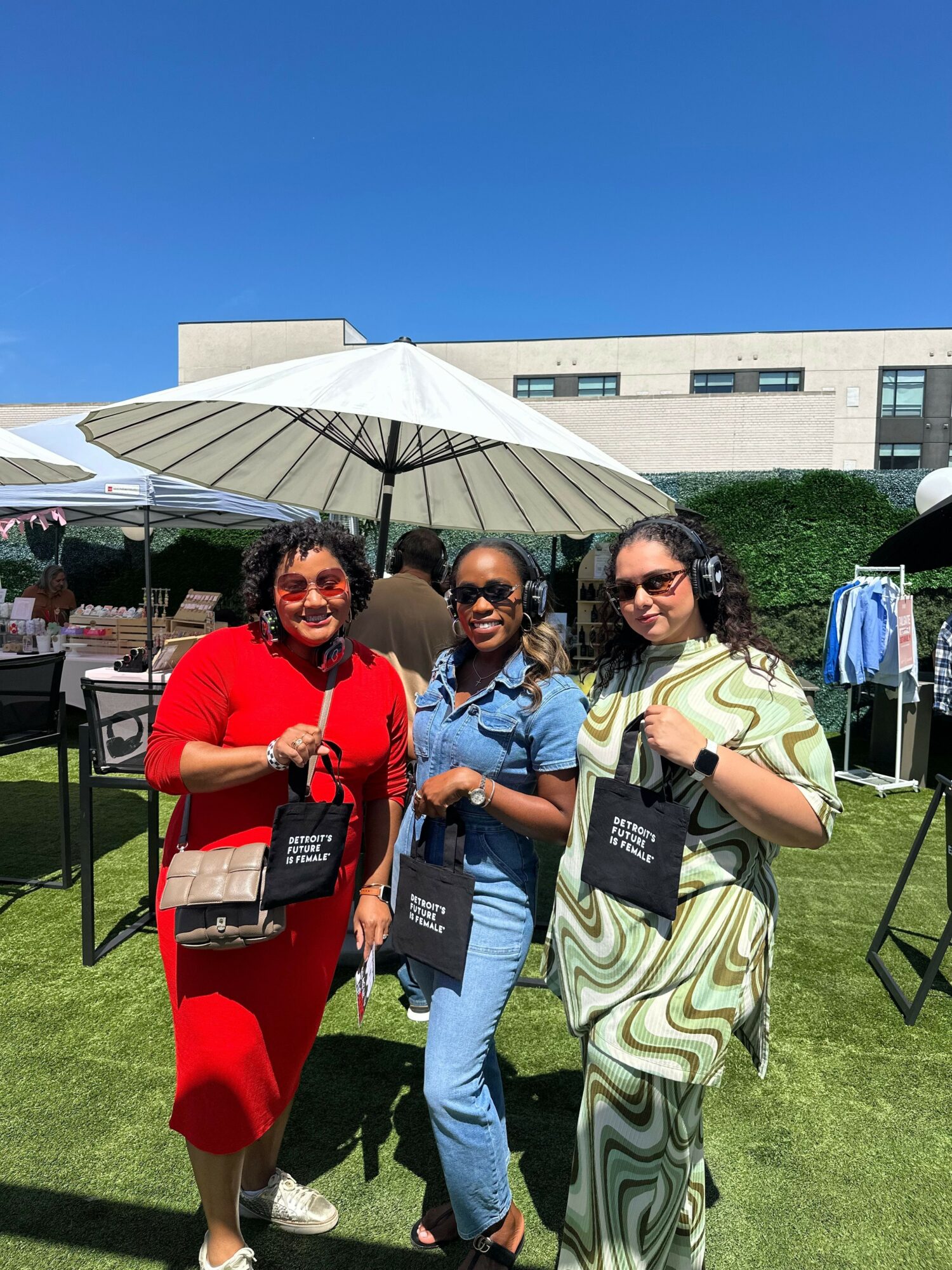 Three women standing outdoors under an umbrella, holding black books, on a sunny day with a building in the background.