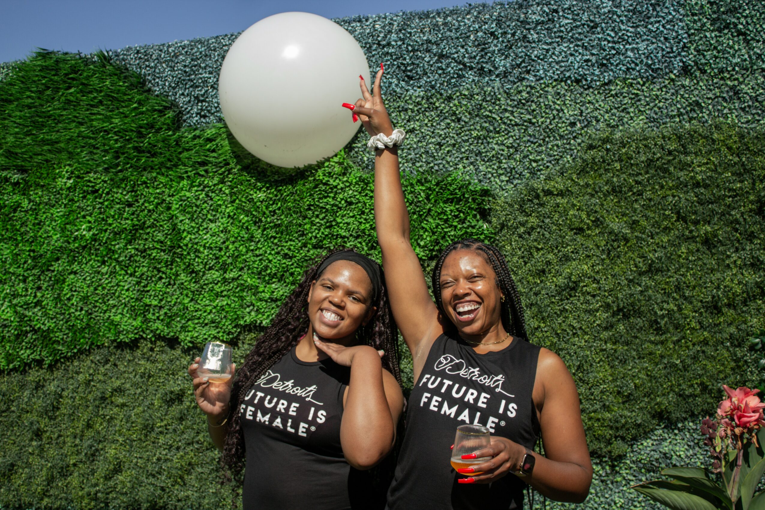 Two women smiling, one holding a drink, the other reaching up towards a large white ball, standing in front of a green wall.
