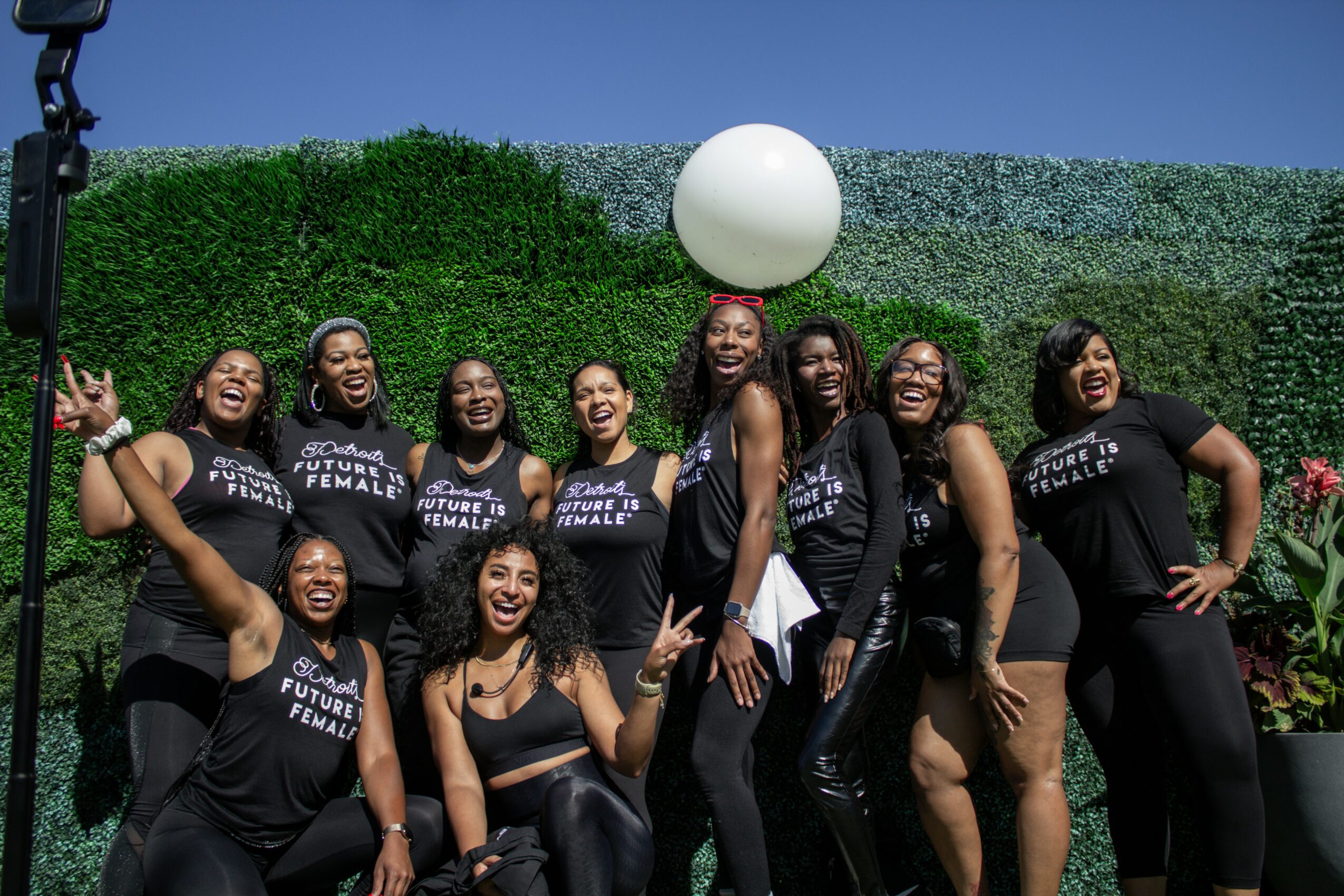 Group of ten women smiling and posing outdoors in front of a green hedge, wearing matching black T-shirts, some with arms raised.