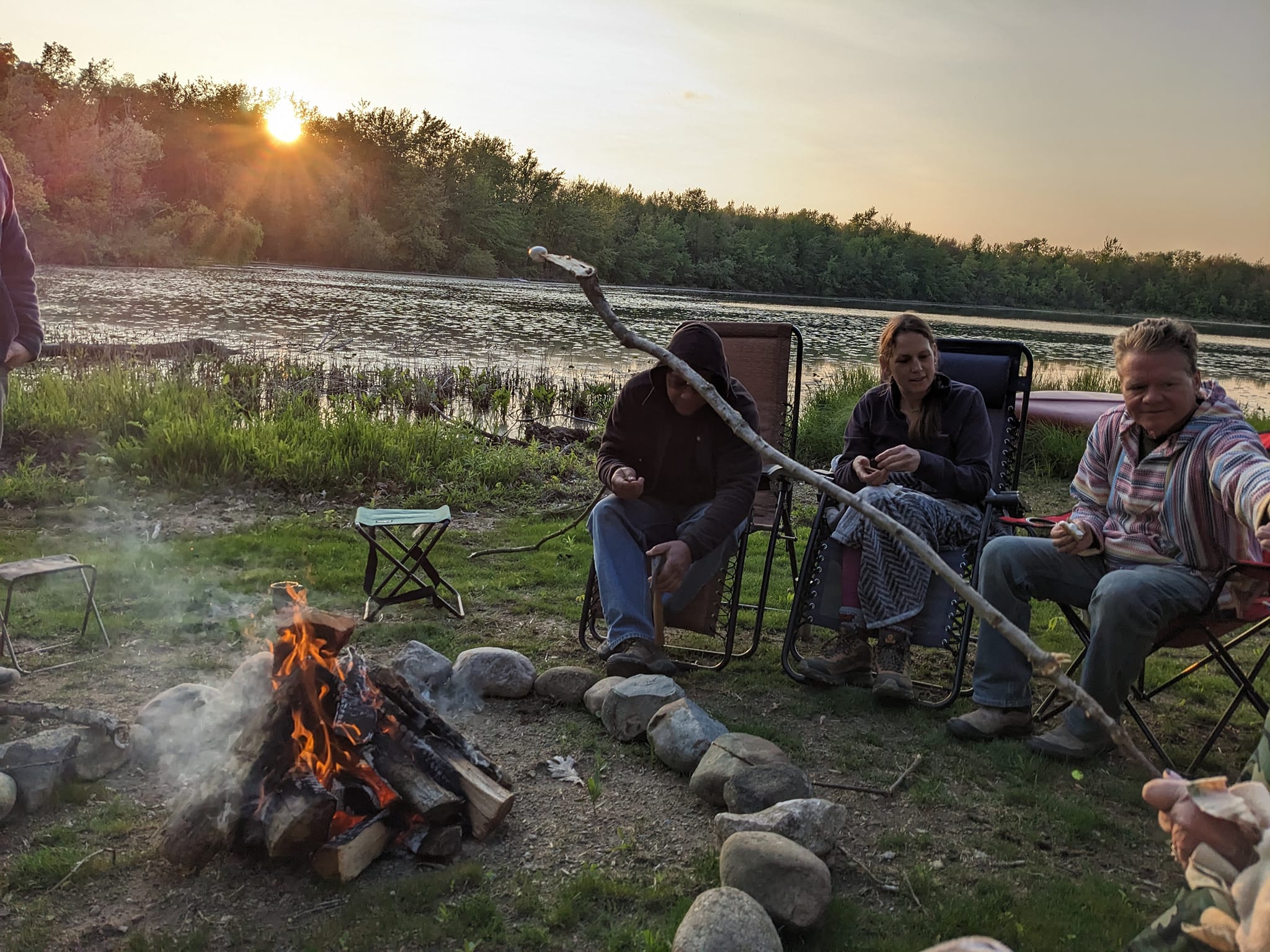 Three people sit near a campfire outdoors with a river and trees in the background during sunset.