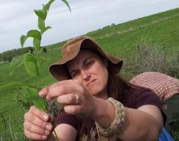 Woman outdoors holding a small plant, with green fields and cloudy sky in background.