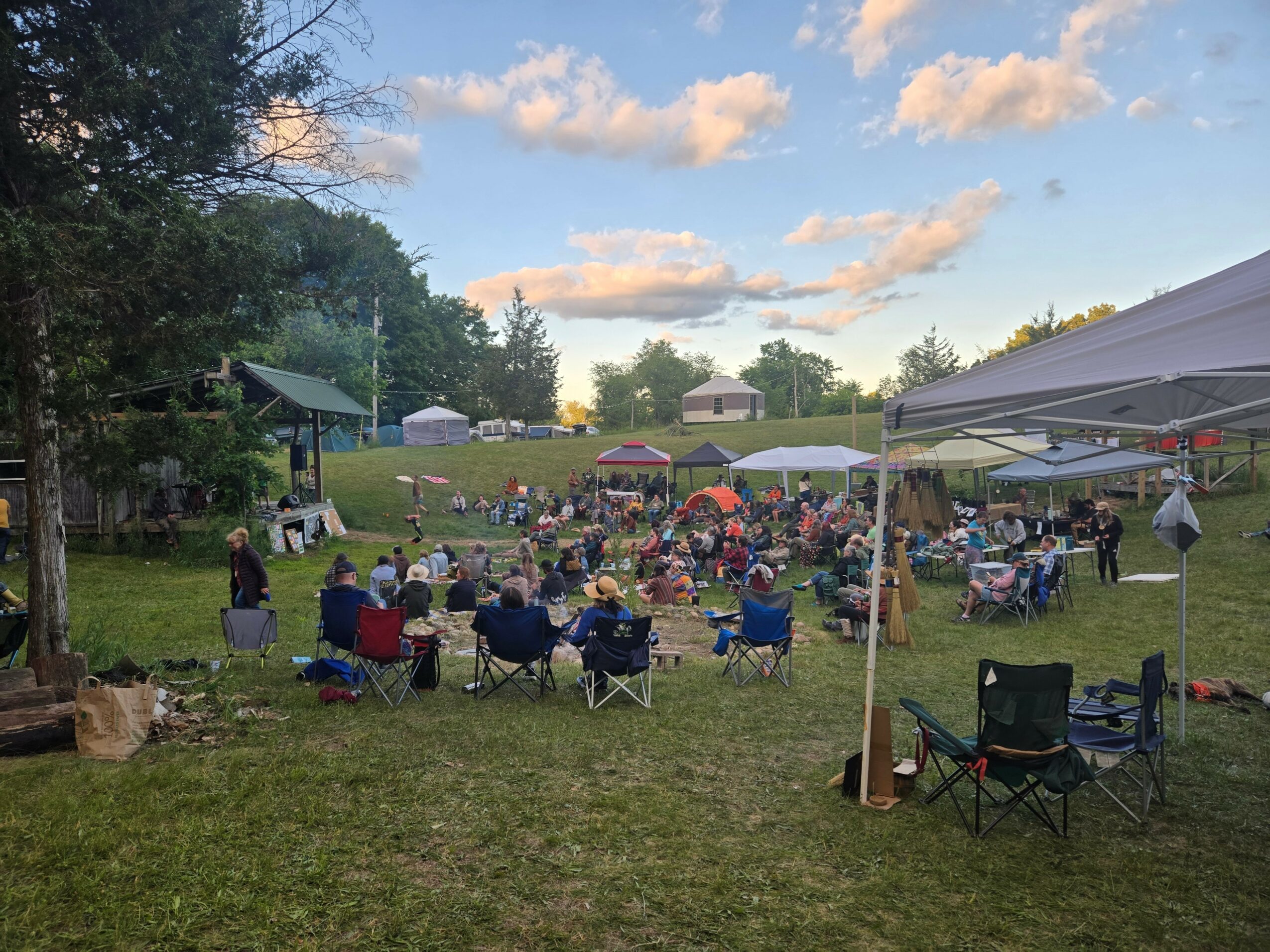 People gathered outdoors on a grassy field with tents and umbrellas, enjoying a sunny day with trees and a blue sky.
