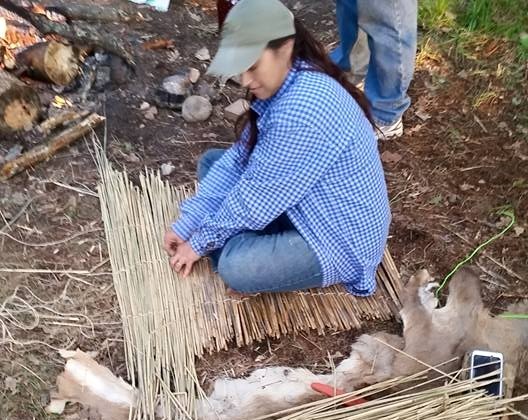 Person sitting on ground with a hat, near goats and a small fence, outdoors in a natural setting.