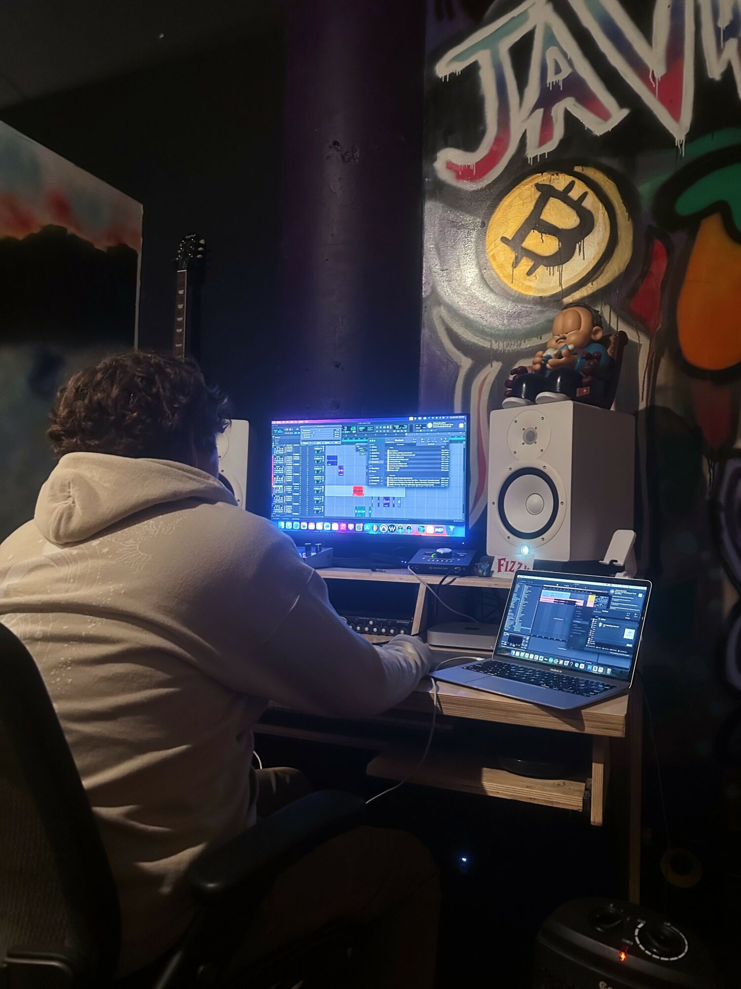 Person with curly hair wearing a hoodie working on a music production computer setup in a dark room with graffiti art, including a Bitcoin symbol.